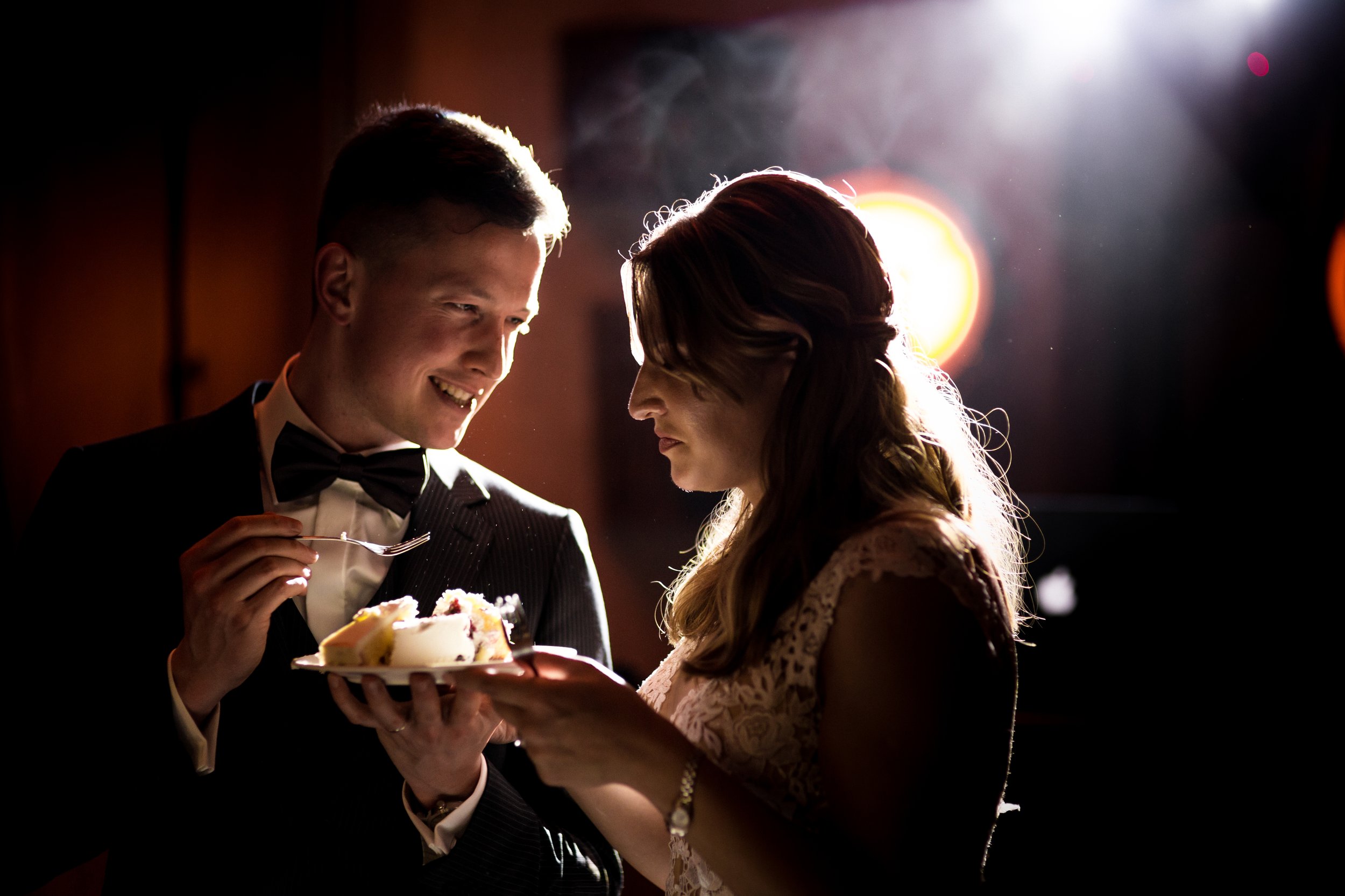 A man in a tuxedo offering a plate of cake to a woman in a lace dress at a dimly lit event, with bright backlighting.