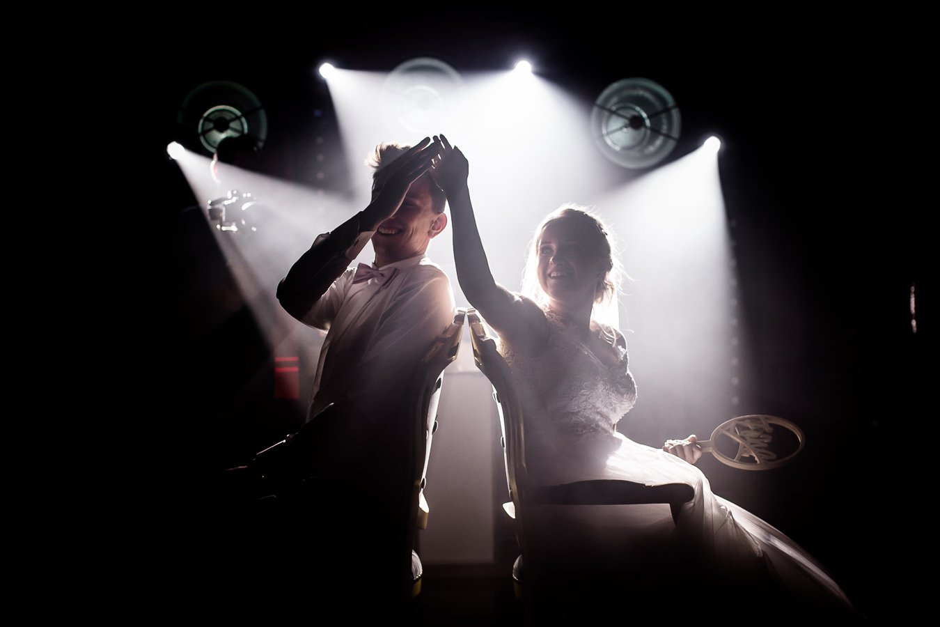Two people sitting back-to-back on chairs on stage, smiling and touching hands, surrounded by bright stage lights.