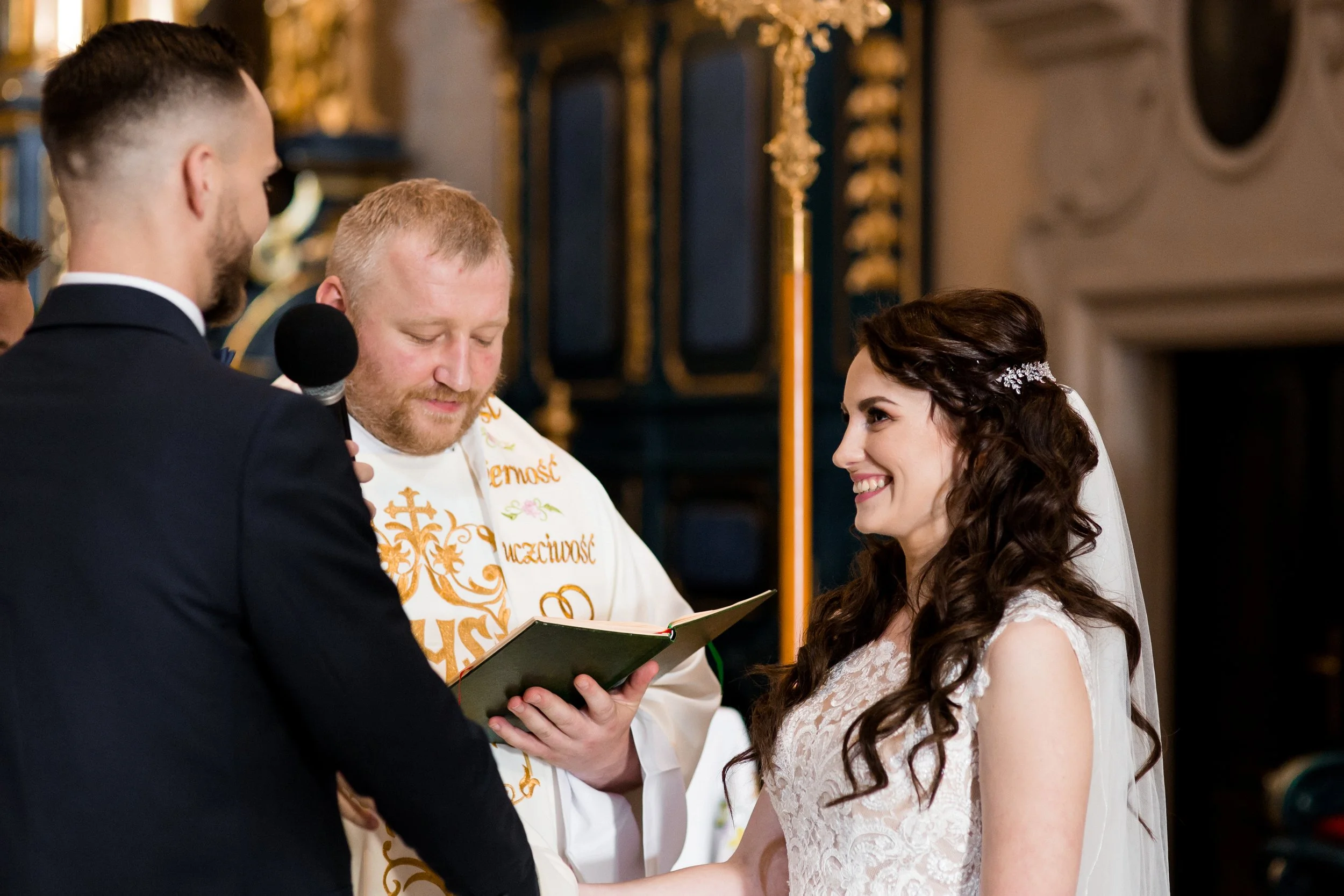A bride and groom holding hands during their wedding ceremony in a church, with a priest reading from a book. The bride has long, dark, curly hair and is wearing a white lace wedding dress, while the groom has short hair and is dressed in a black sui