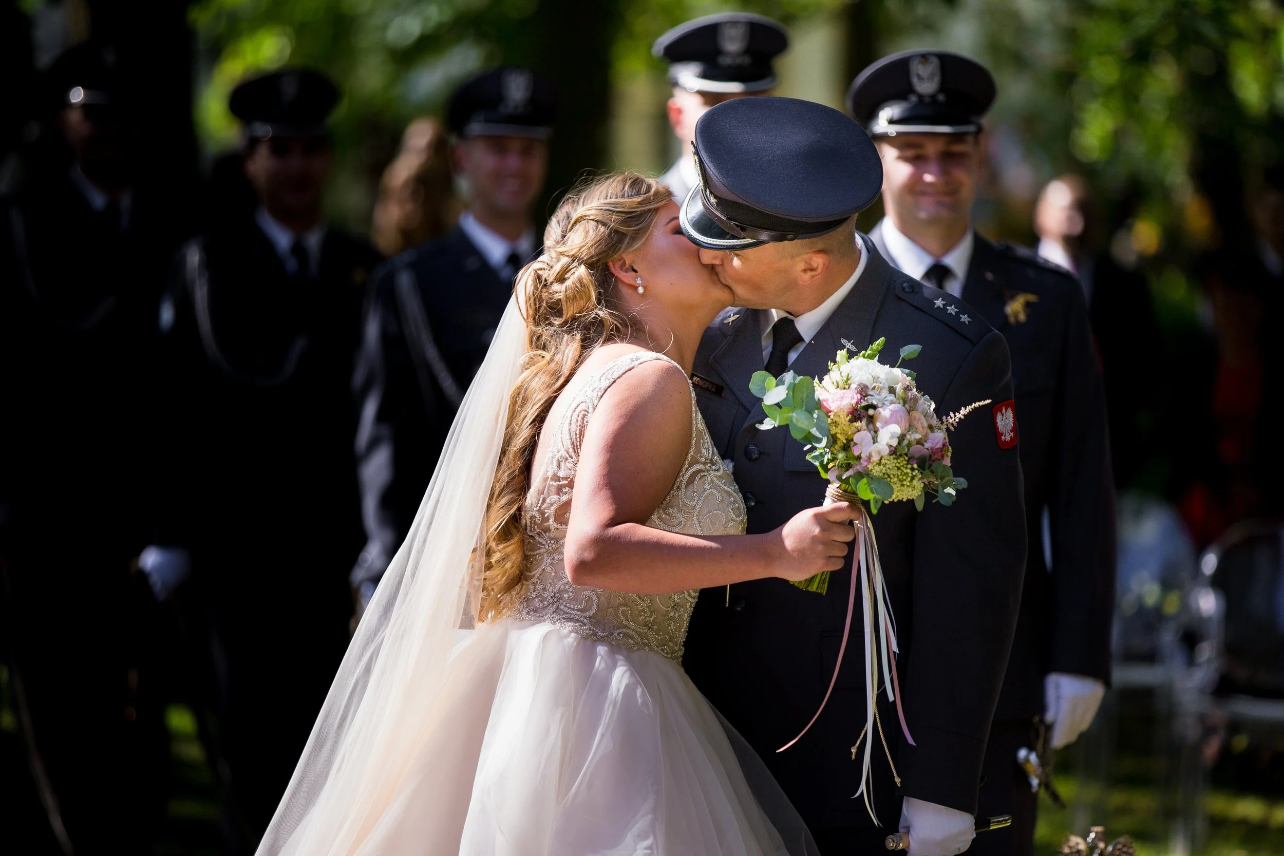 A bride and a groom in military uniform kissing at an outdoor wedding ceremony, with bridesmaids and groomsmen in the background.