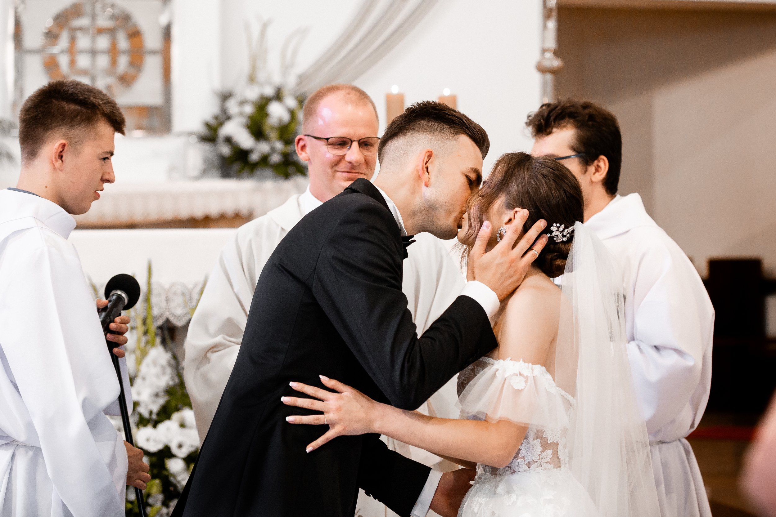 A couple kissing during a wedding ceremony, surrounded by officiants and a person in a white robe holding a microphone.