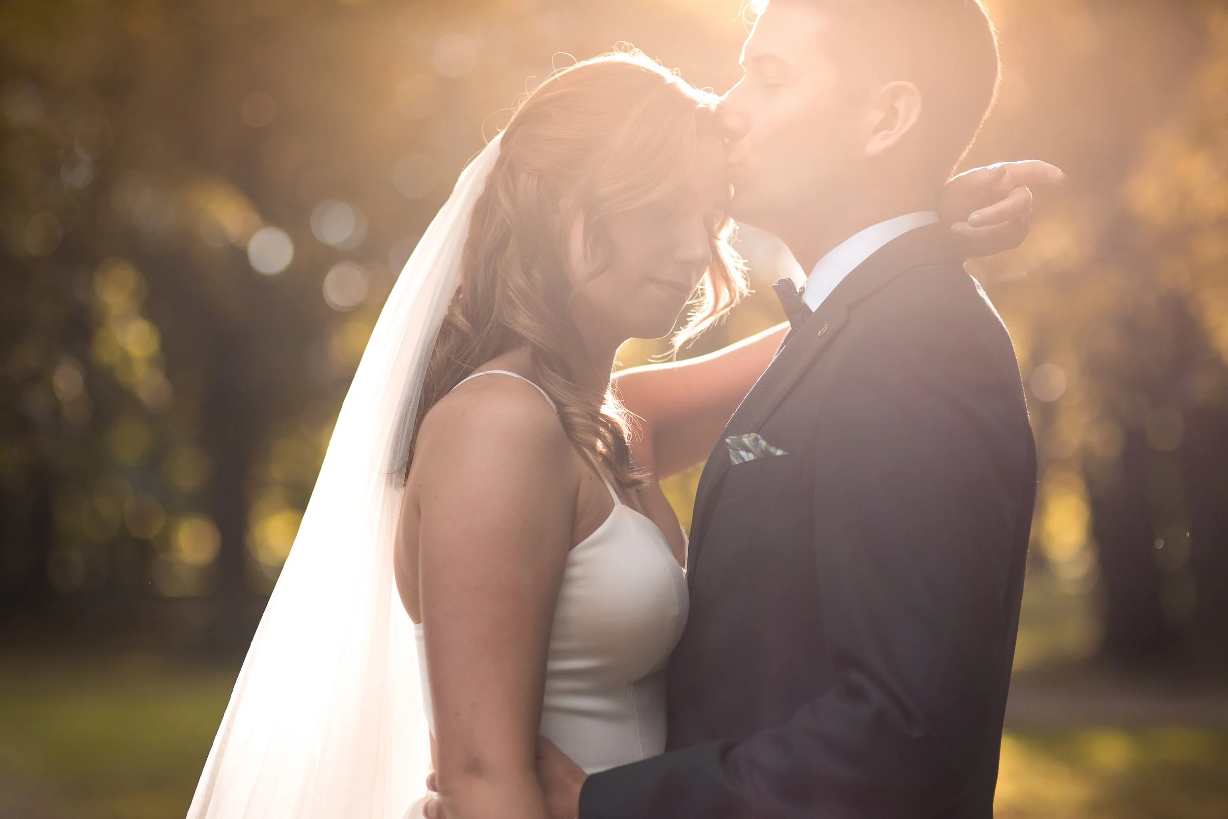 Bride and groom sharing a kiss outdoors during sunset, with sunlight backlighting them and blurred trees in the background.