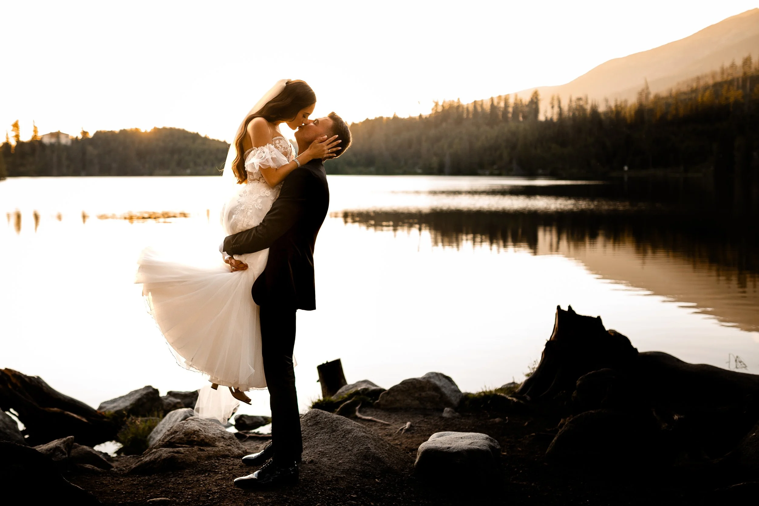 A couple dressed in wedding attire sharing a kiss by a lake during sunset, with mountains and trees in the background.