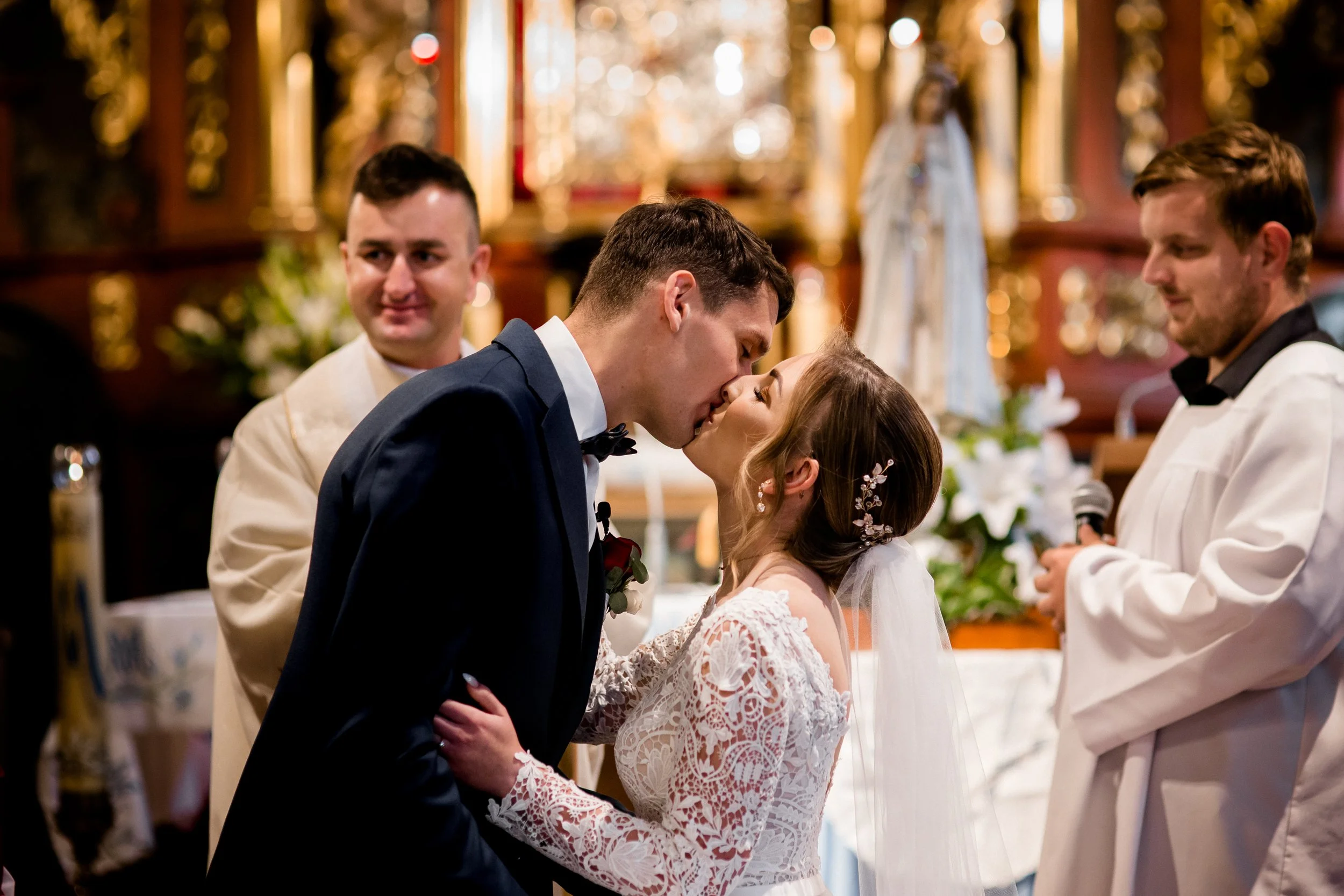 A bride and groom kiss during their wedding ceremony in a church, with two officiants or witnesses observing in the background.