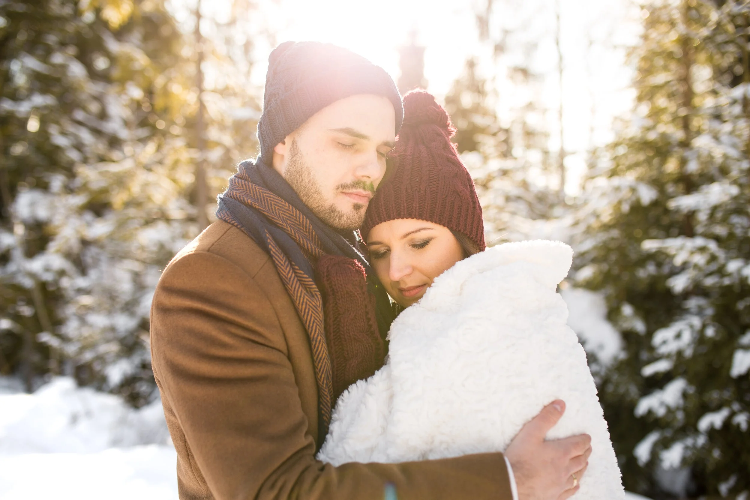 A couple embracing in a snowy forest, wearing warm clothing and knit hats, with sunlight shining through trees.