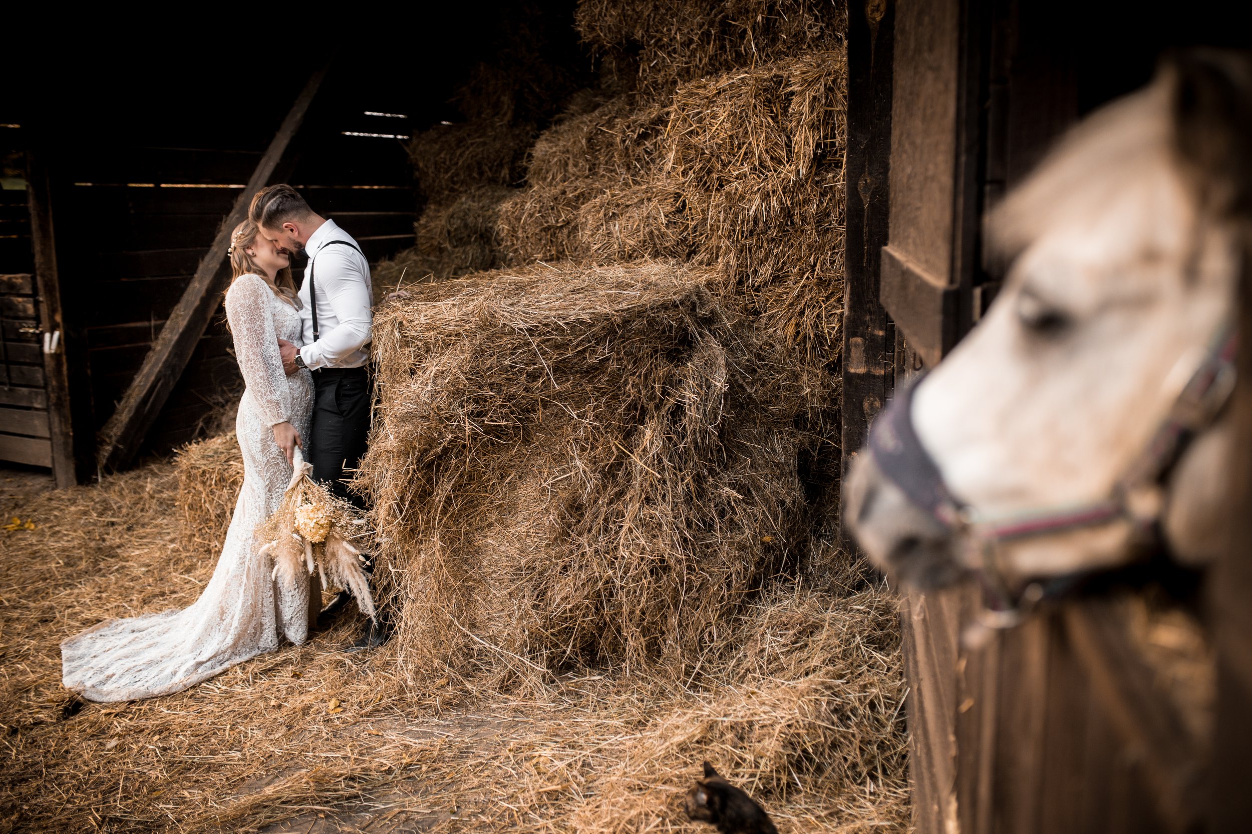 Panna młoda i pan młody stoją blisko siebie w rustykalnej stodole otoczonej sianem, po prawej stronie częściowo widoczny jest koń.