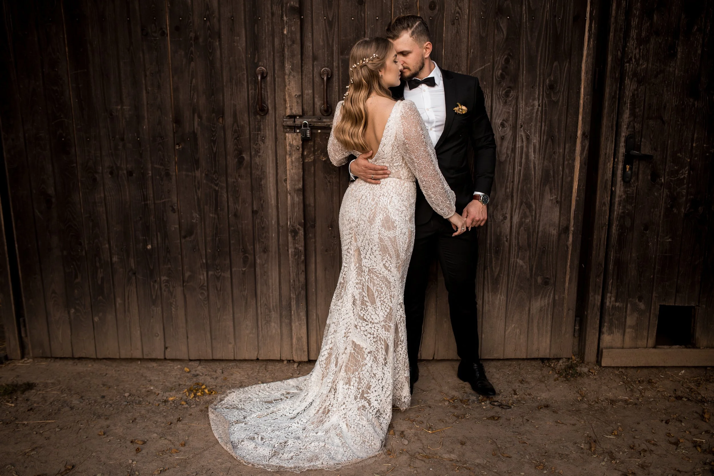A bride and groom standing close together in front of a wooden wall, embracing and touching foreheads. The bride is wearing a long, lace wedding dress, and the groom is dressed in a black suit with a bow tie.