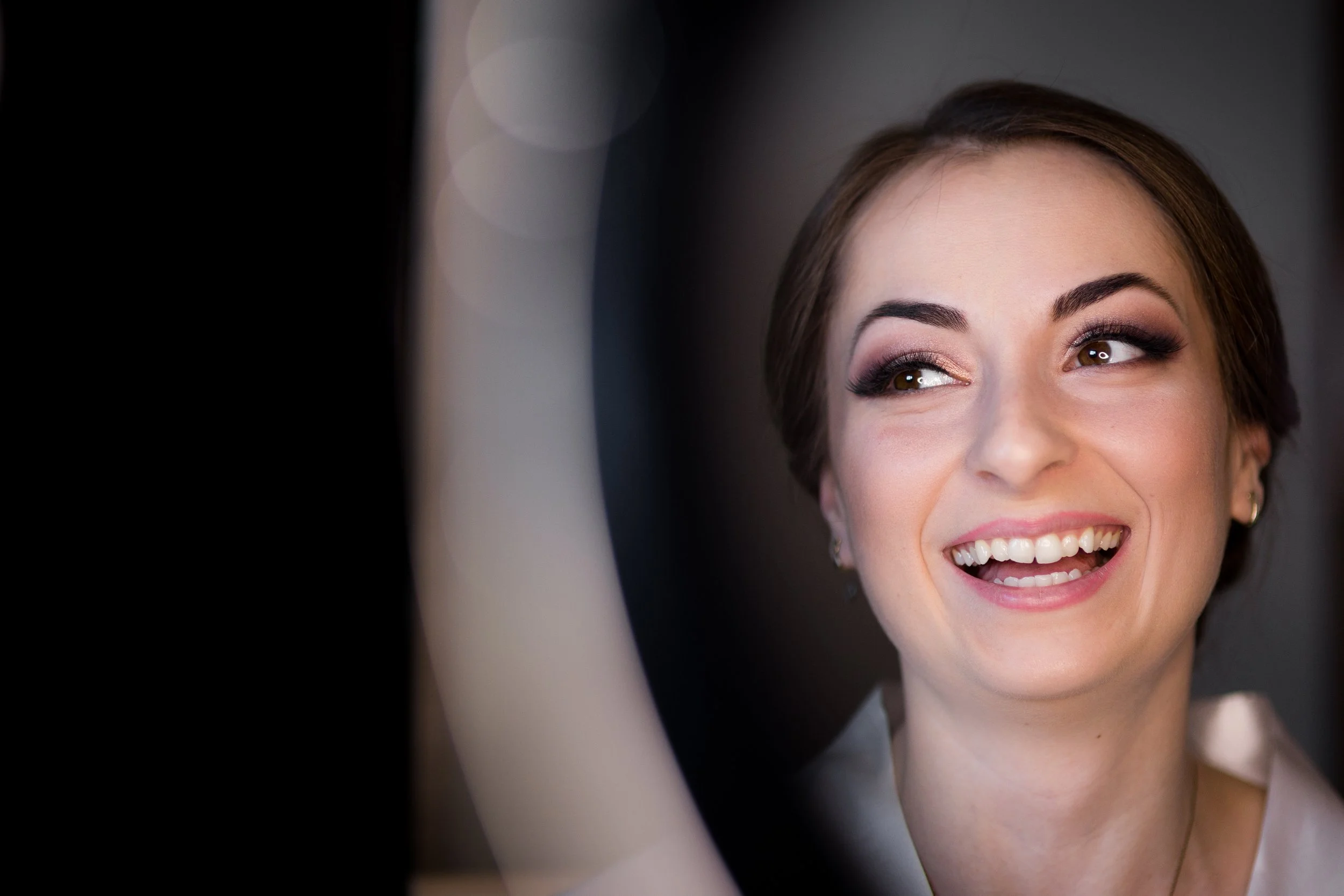 A woman smiling and looking to the side while applying makeup or adjusting her hair, with a blurred background.