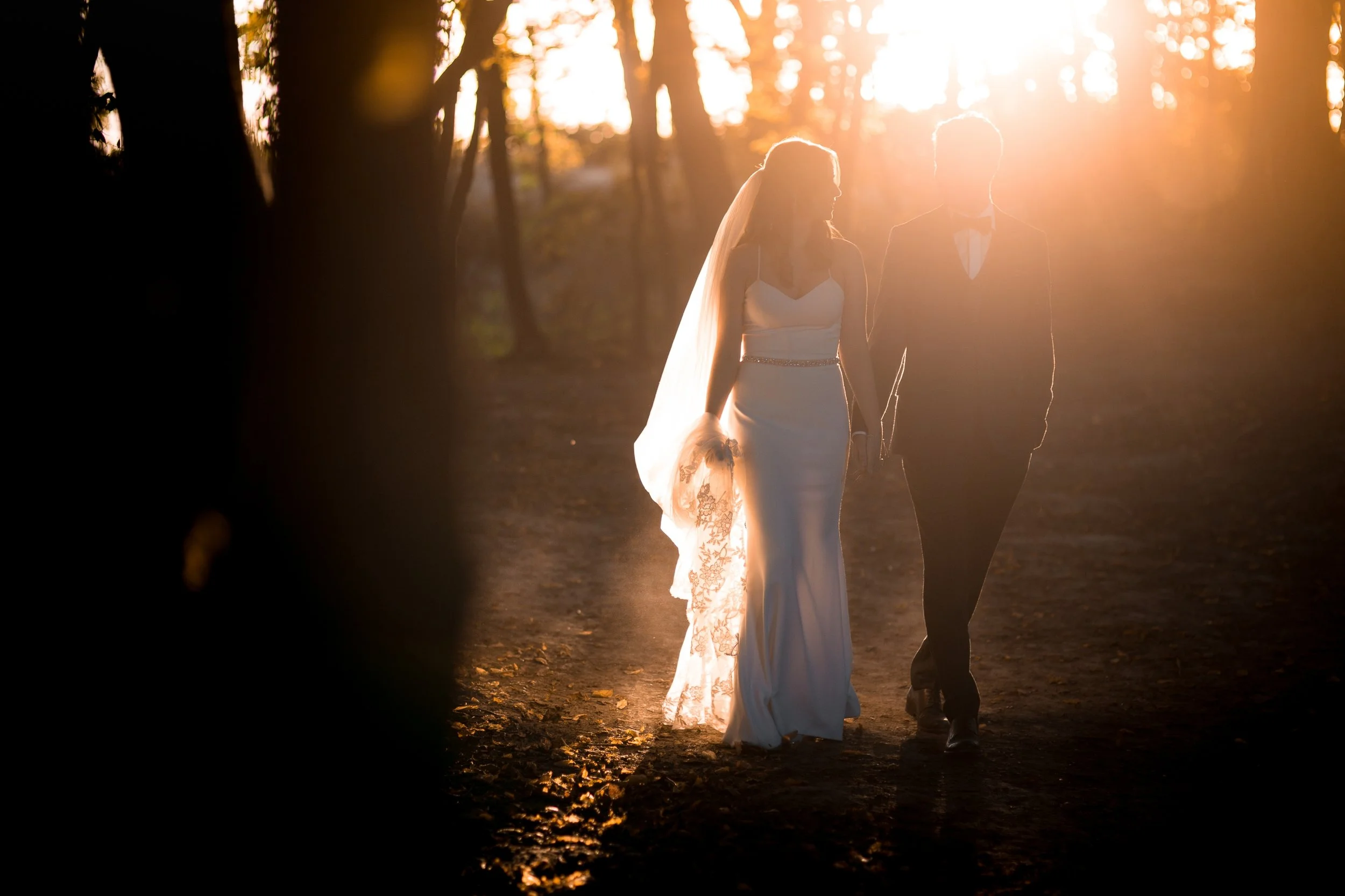 Silhouetted bride and groom walking hand in hand in a wooded area during sunset.