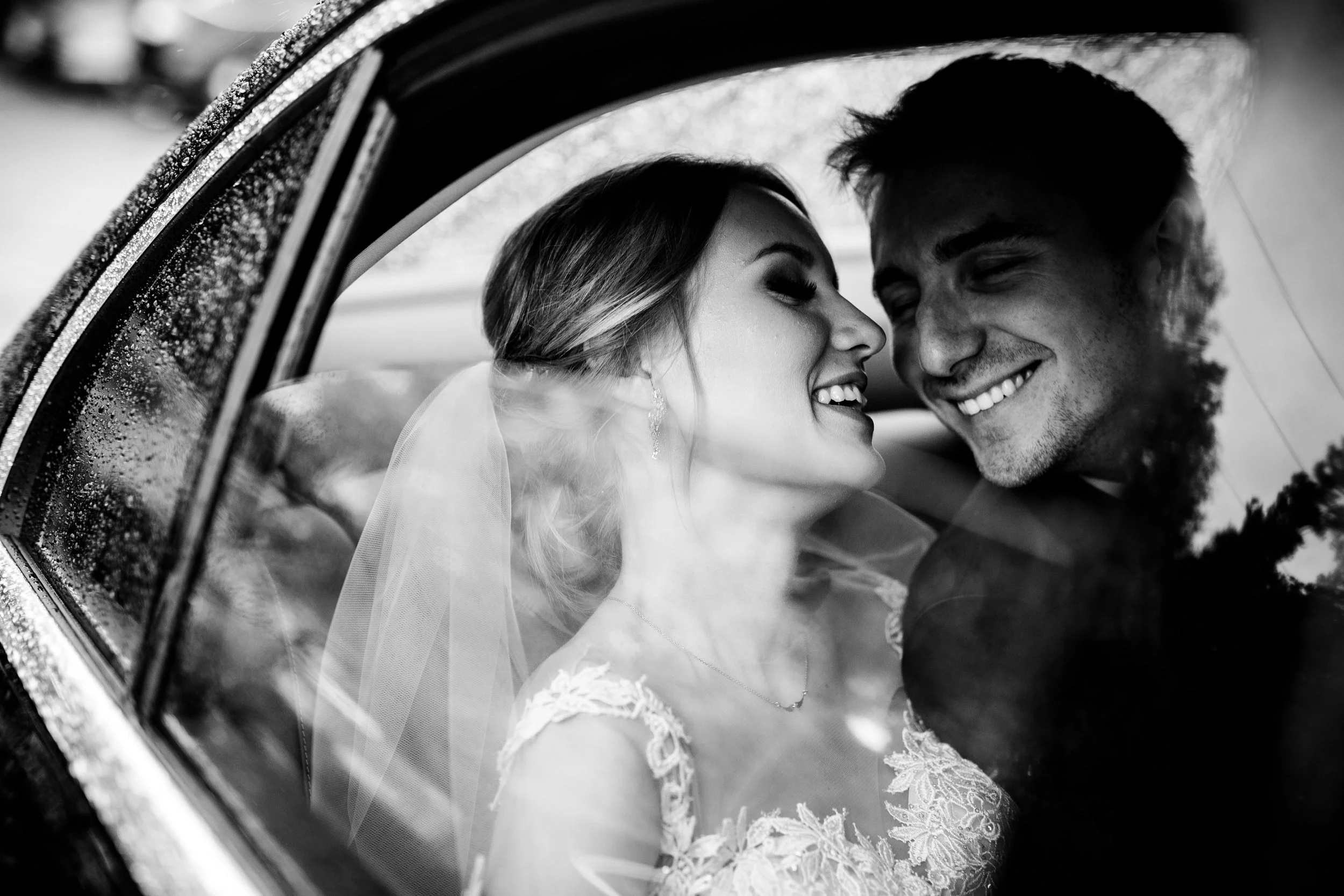 Czarno-białe zdjęcie uśmiechniętej pary młodej w samochodzie, na szybie widać krople deszczu.
