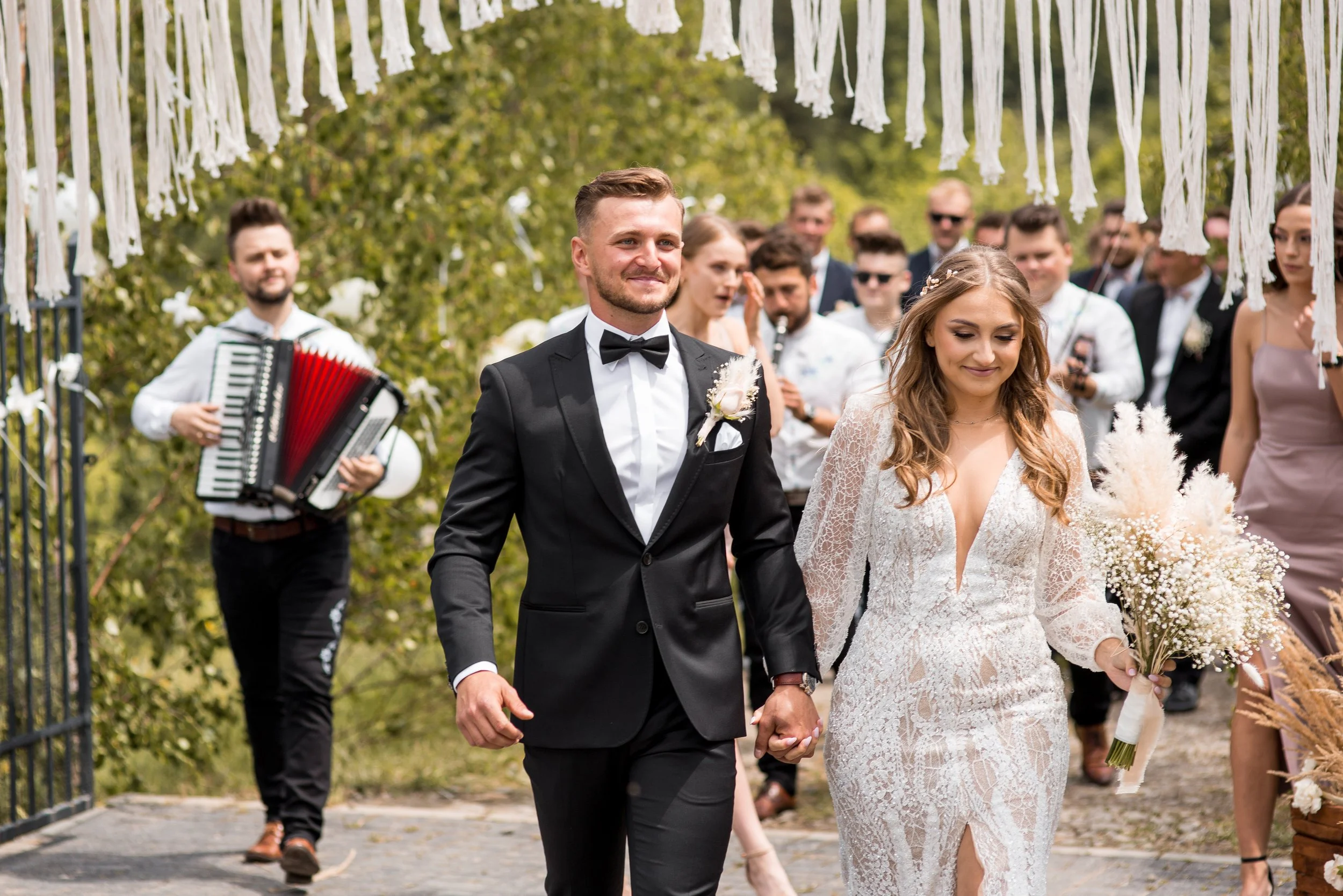 A bride and groom holding hands and walking outdoors during their wedding ceremony, surrounded by family and friends, with a musician playing an accordion in the background, under a decorated white canopy.