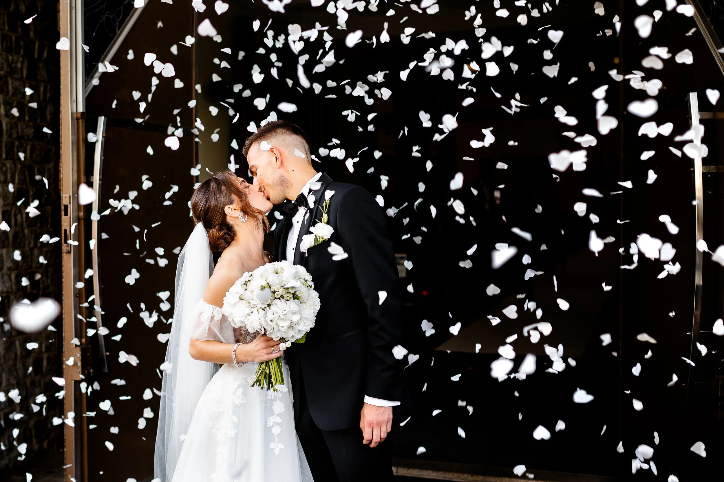 Bride and groom sharing a kiss surrounded by white confetti, holding a bouquet of white flowers, in a wedding celebration.