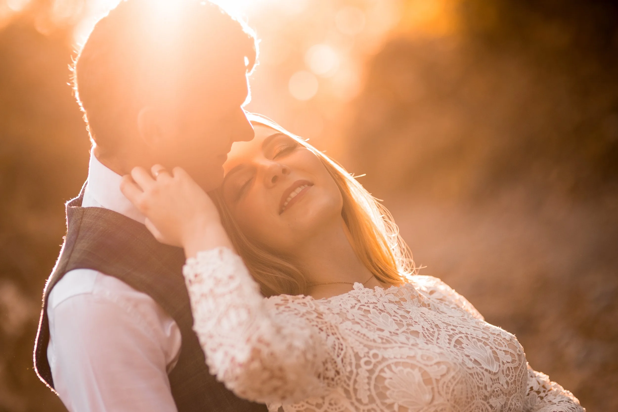 A couple embraces during sunset, with the woman smiling and the man's face partially in shadow.