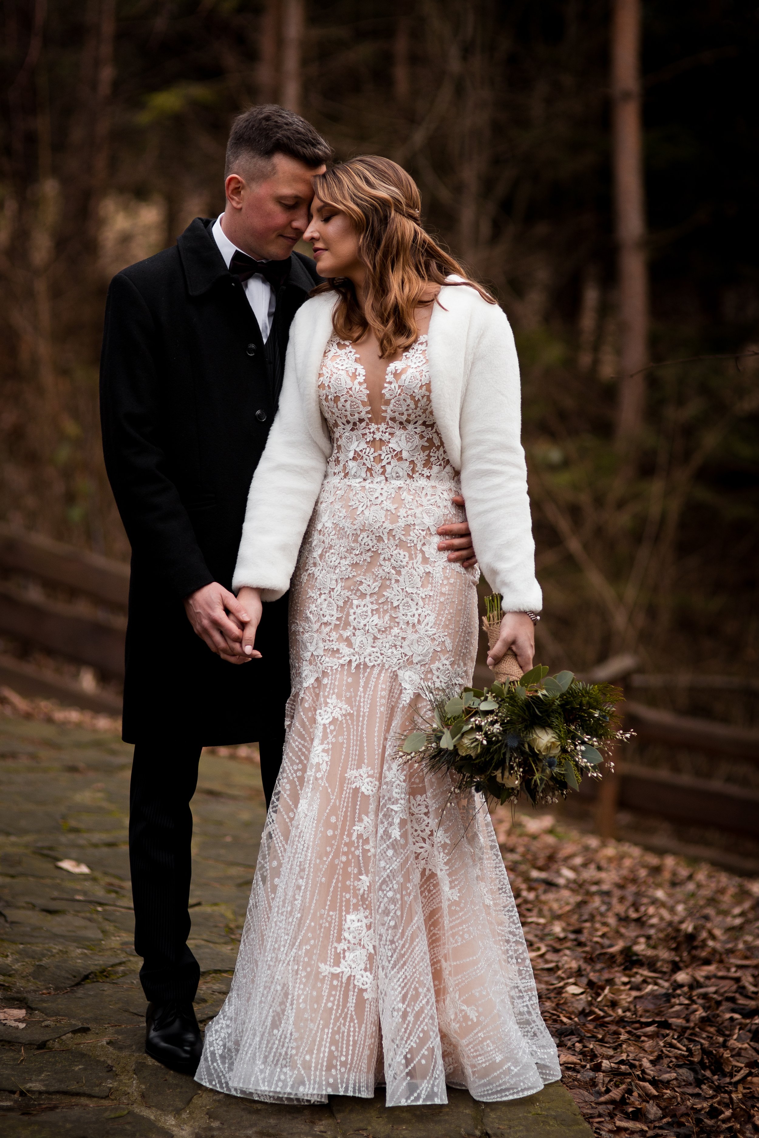 A couple dressed in wedding attire walking hand in hand outdoors in a wooded area, with the bride holding a bouquet of flowers.