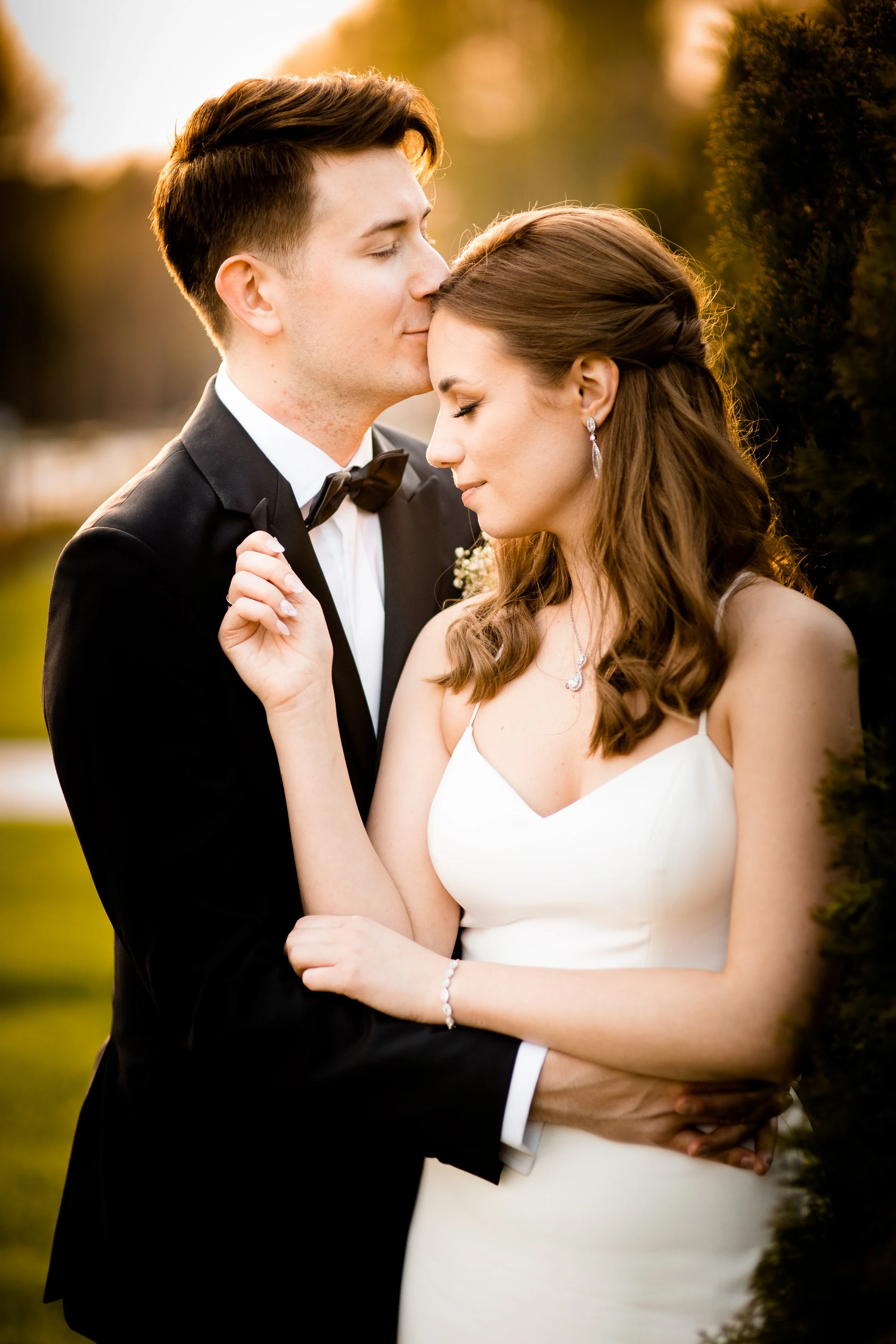 A newlywed couple standing close together outdoors during sunset, with the groom in a black tuxedo and the bride in a white wedding dress, embracing lovingly.