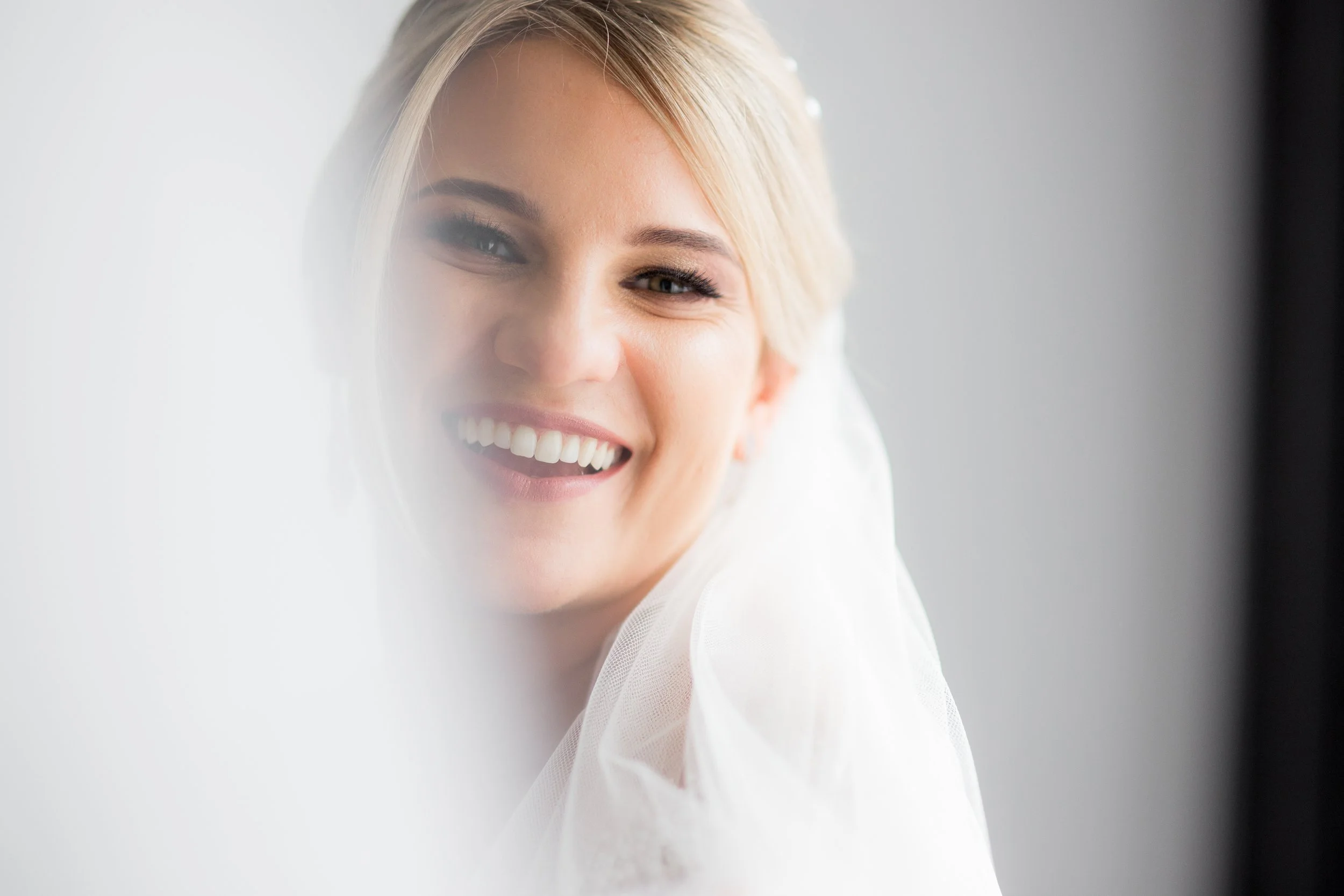 A smiling woman with blonde hair, natural makeup, and white teeth looking at the camera, with a soft light background.