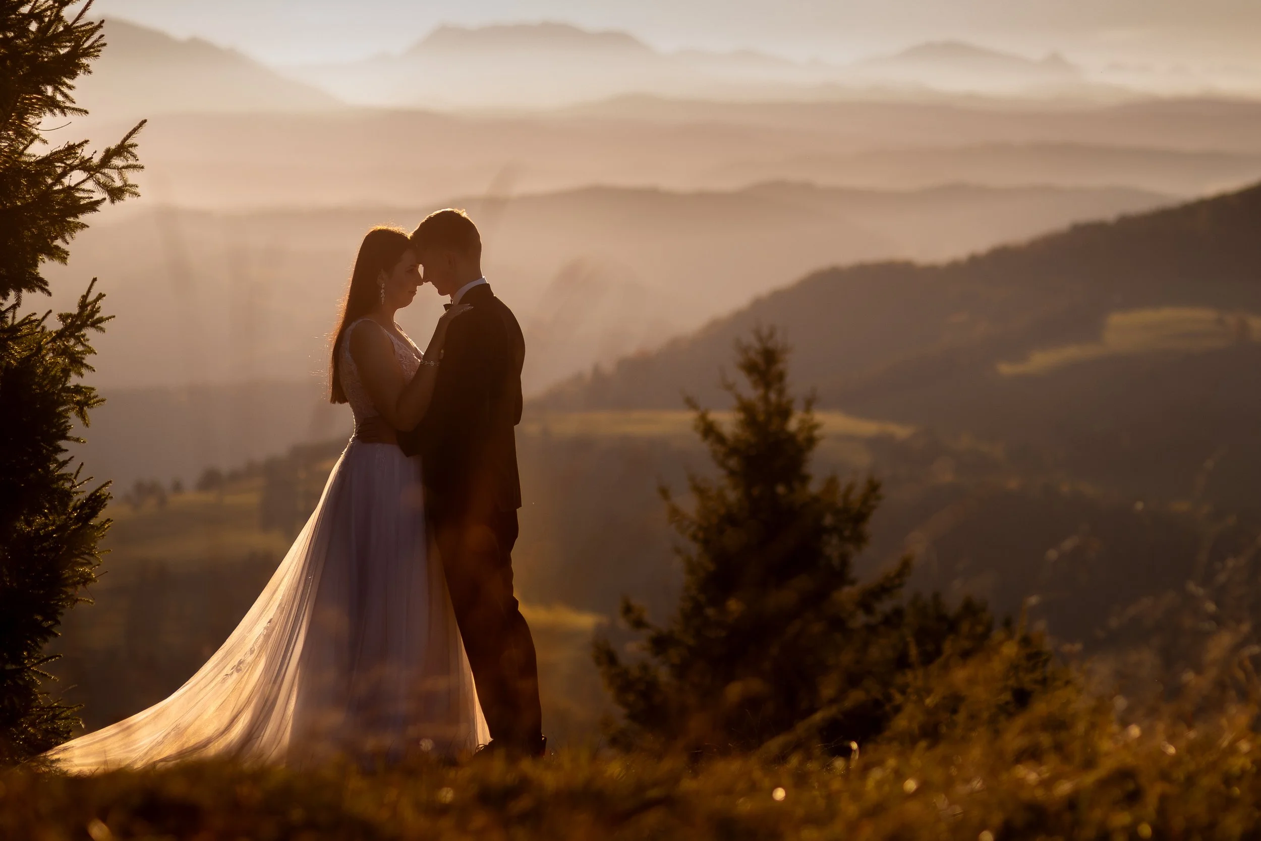Sylwetka panny młodej i pana młodego obejmujących się na zboczu wzgórza w Pieninach o zachodzie słońca, z górami i drzewami i Tatrami w tle.
