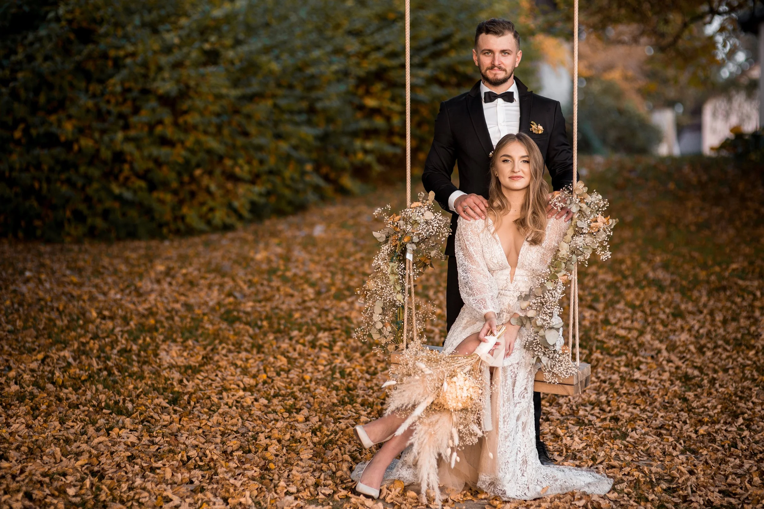 Para młoda pozująca na świeżym powietrzu jesienią. Panna młoda siedzi na udekorowanej huśtawce, a pan młody stoi za nią.