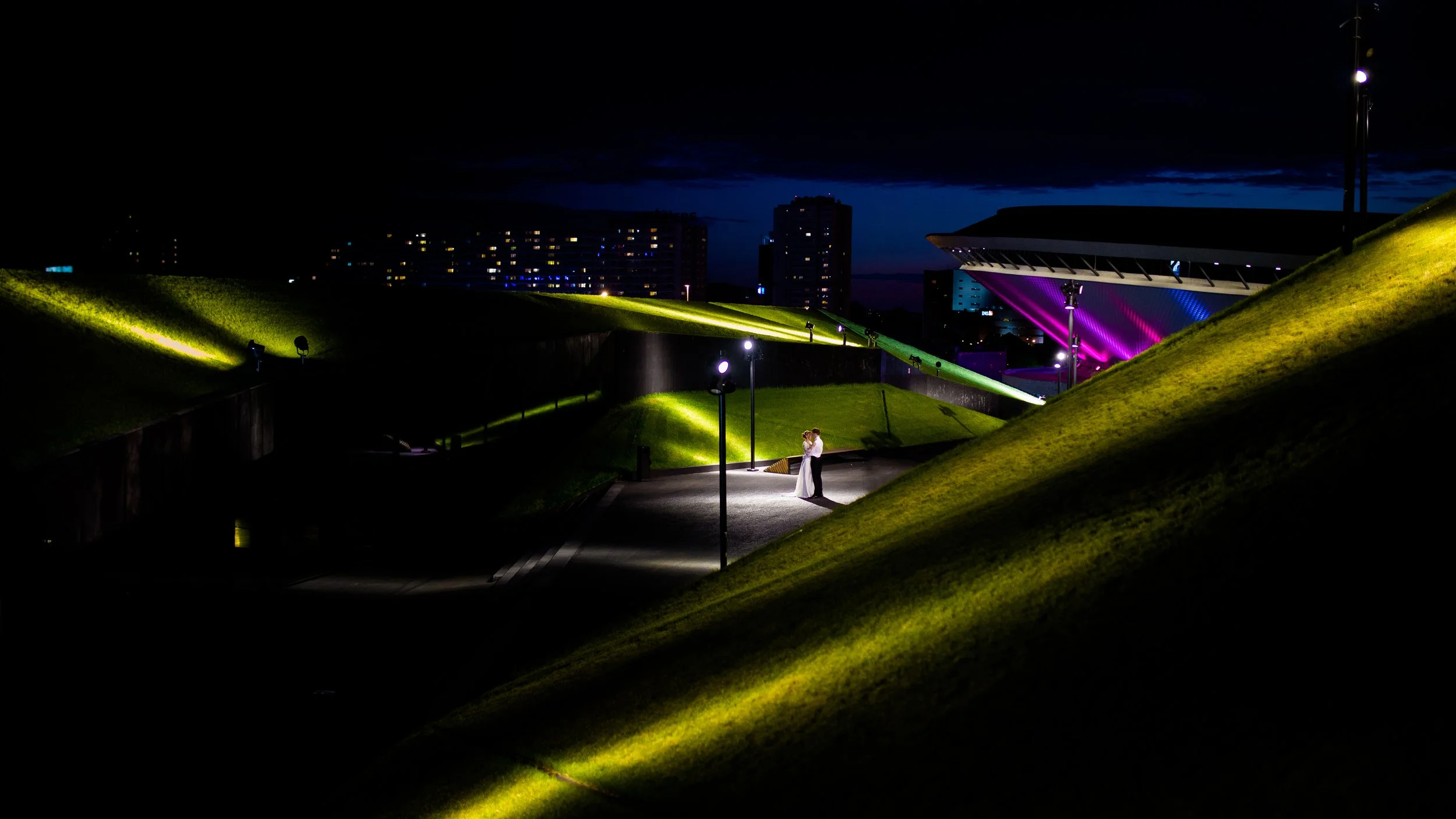 A couple is sharing a kiss under a lamppost on a small plaza at night, with green grassy hills, bright lights, and modern buildings in the background.