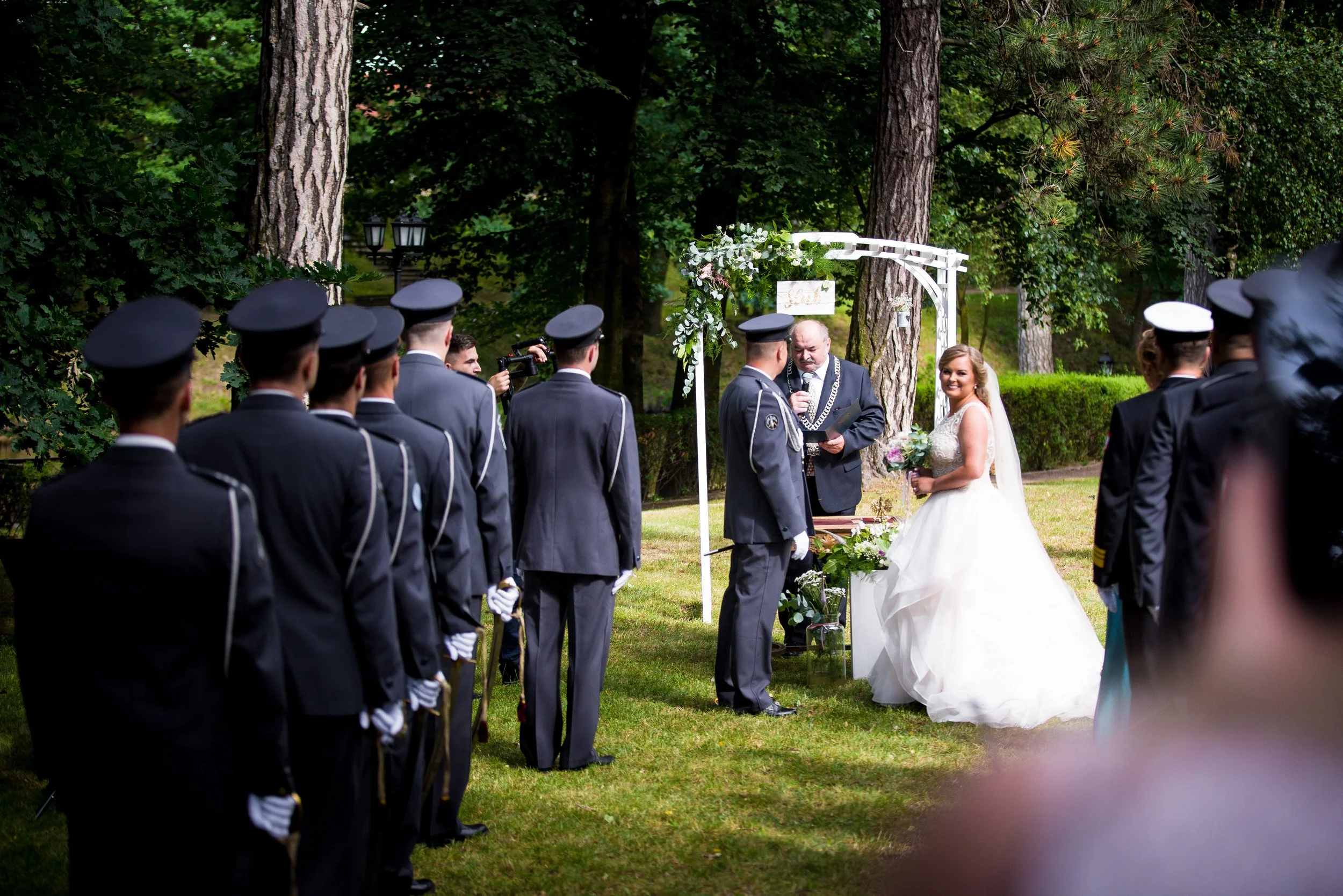 A wedding ceremony outdoors in a forested area with the bride in a white wedding dress holding a bouquet, standing by the groom in a military uniform. A officiant in a dark suit conducts the ceremony under a white arch decorated with flowers. Guests,
