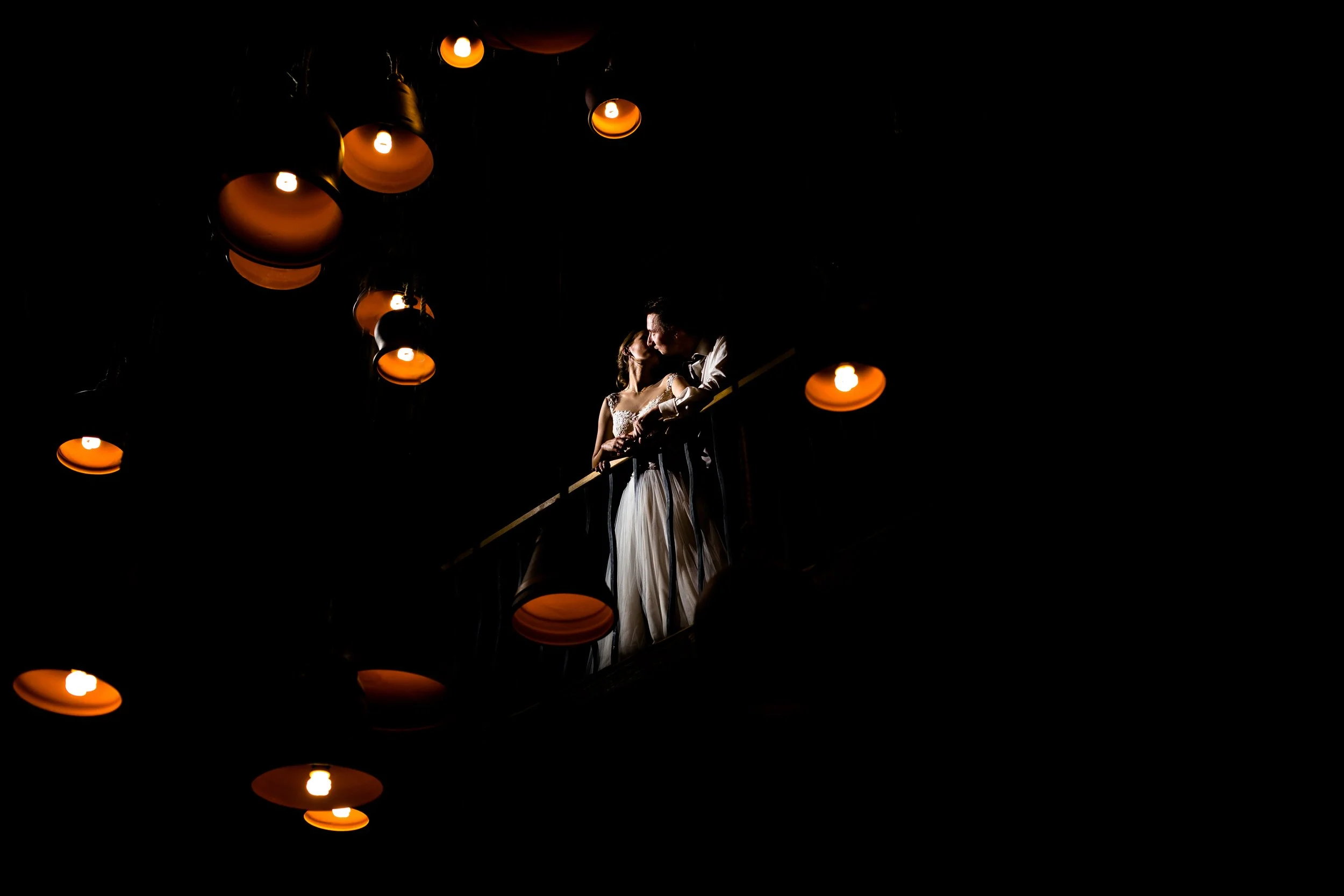Para w strojach ślubnych całuje się na balkonie otoczonym wiszącymi pomarańczowymi lampami, na ciemnym tle.