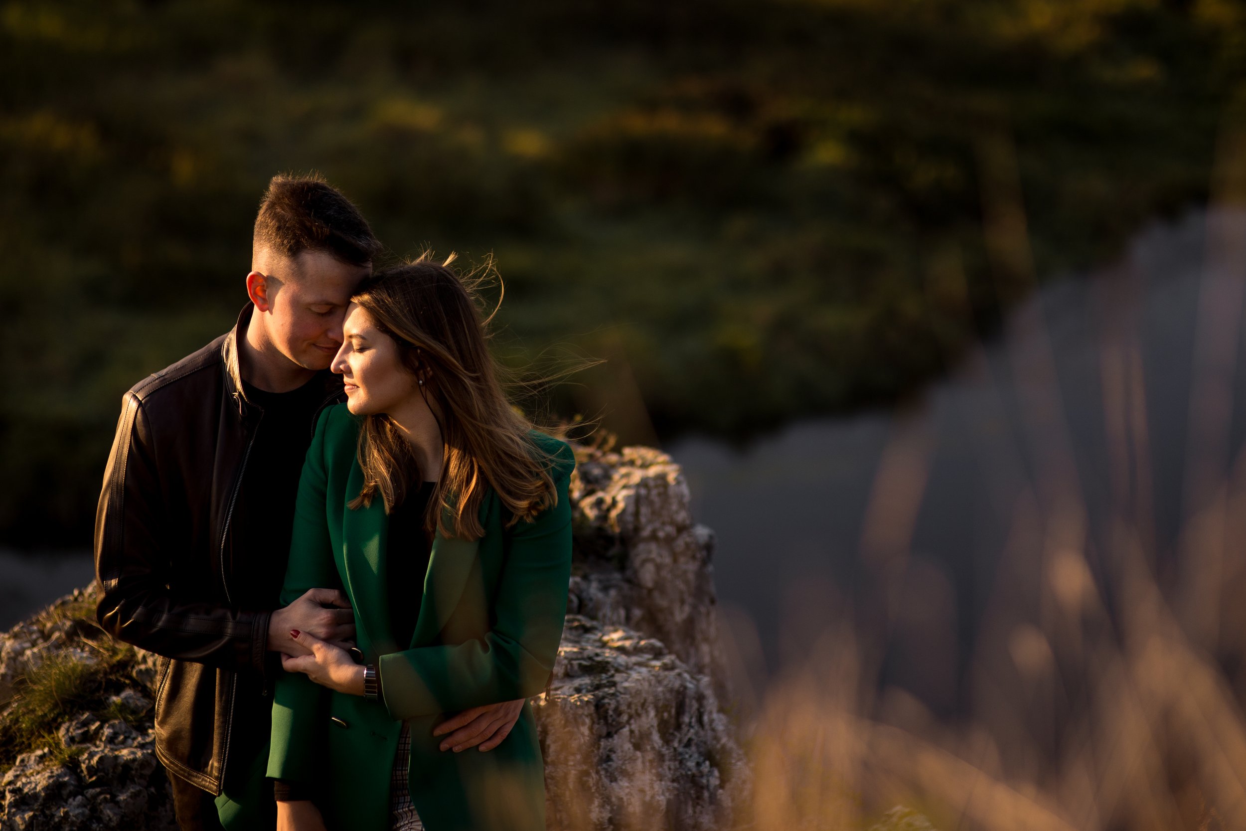 A couple standing close together outdoors during sunset, embracing with their foreheads touching, near a stone structure with a blurred natural background.