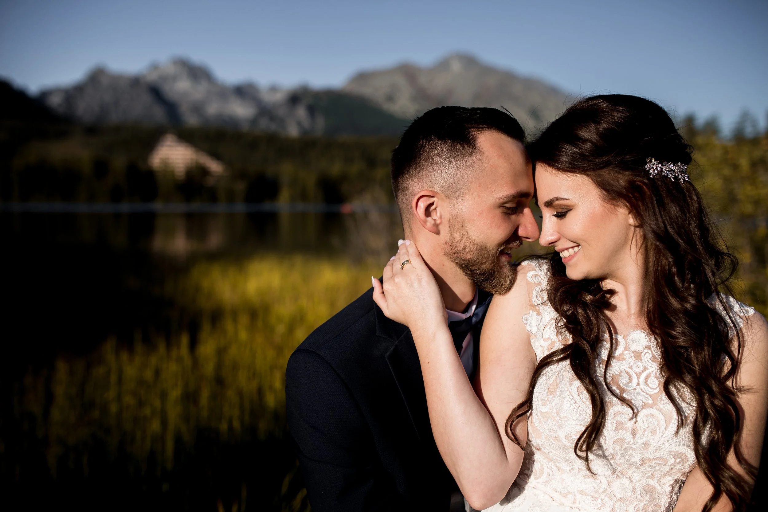 A bride and groom embraced outdoors with mountains and a lake in the background, showing close-up of their faces as they smile and touch foreheads.