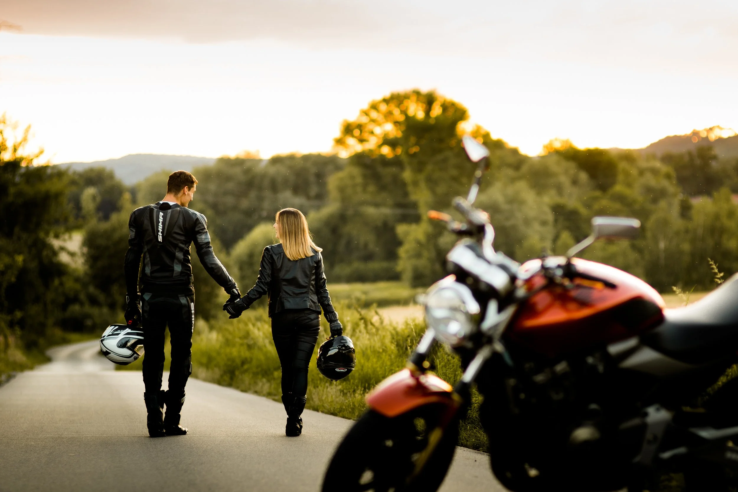 A man and woman in motorcycle gear walking hand-in-hand along a rural road, with two motorcycles parked nearby, during sunset.