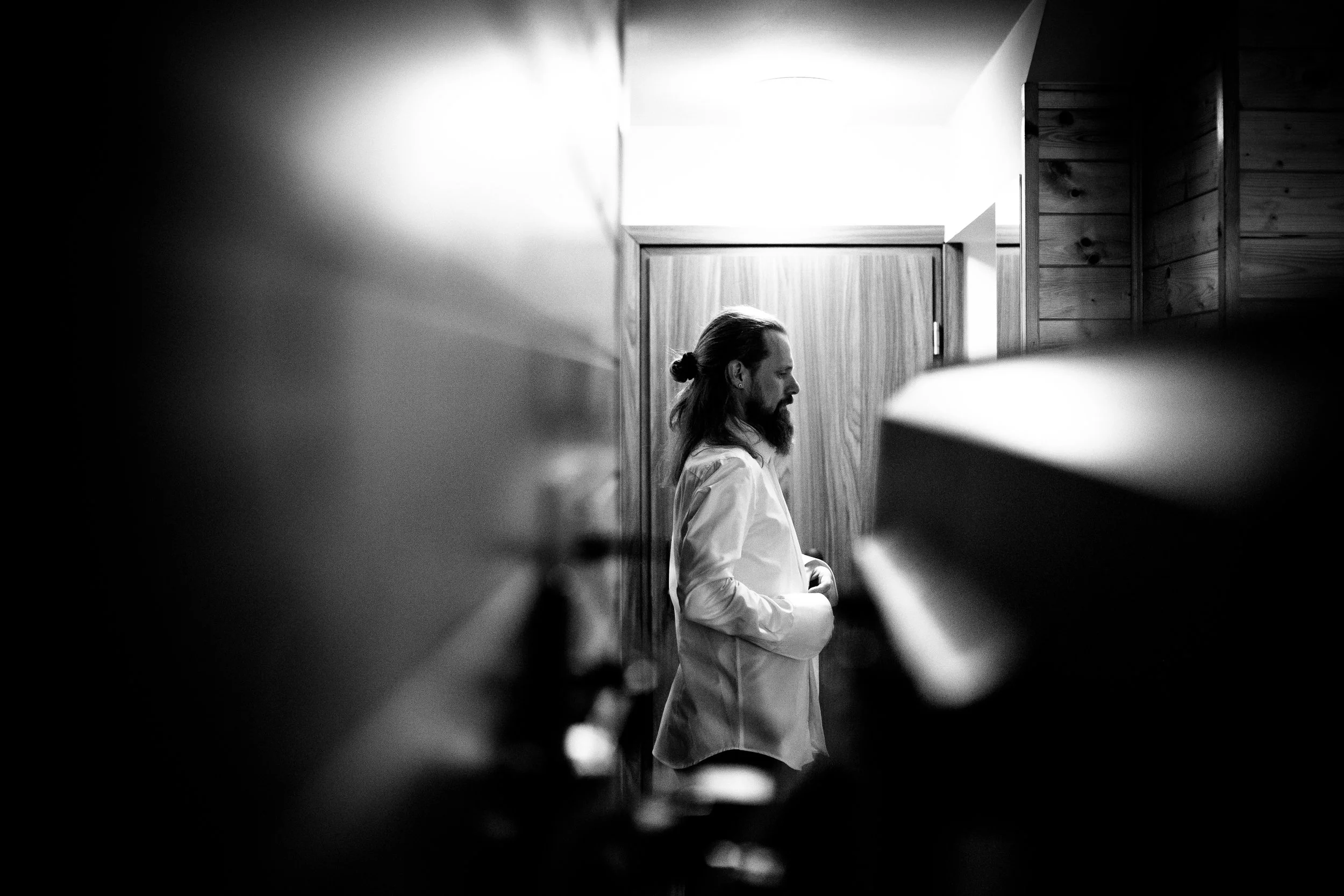 A black and white photo of a man with long hair tied back and a beard, standing in profile with eyes closed, wearing a white shirt, inside a room with wooden paneling.