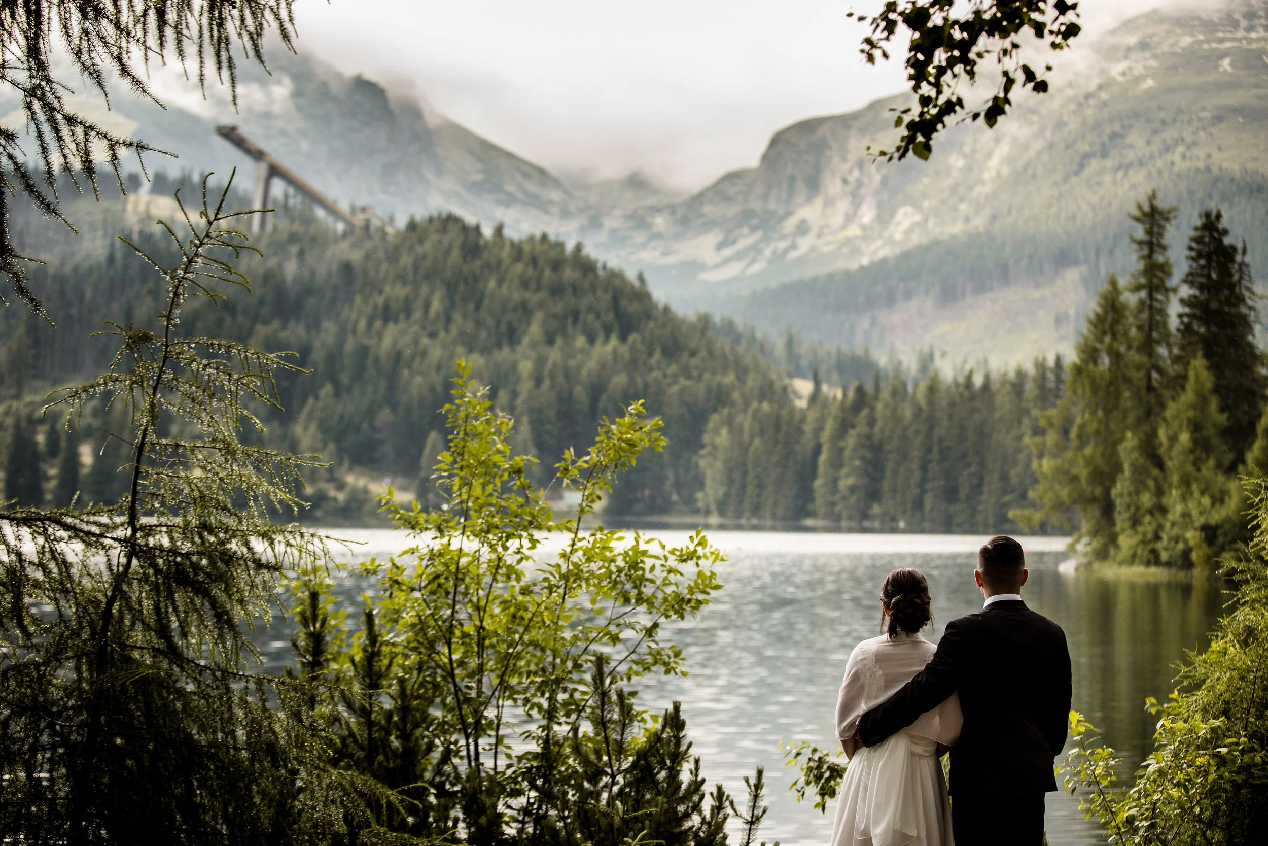 A couple dressed in formal wedding attire standing by a lake, surrounded by trees and mountains in the background.