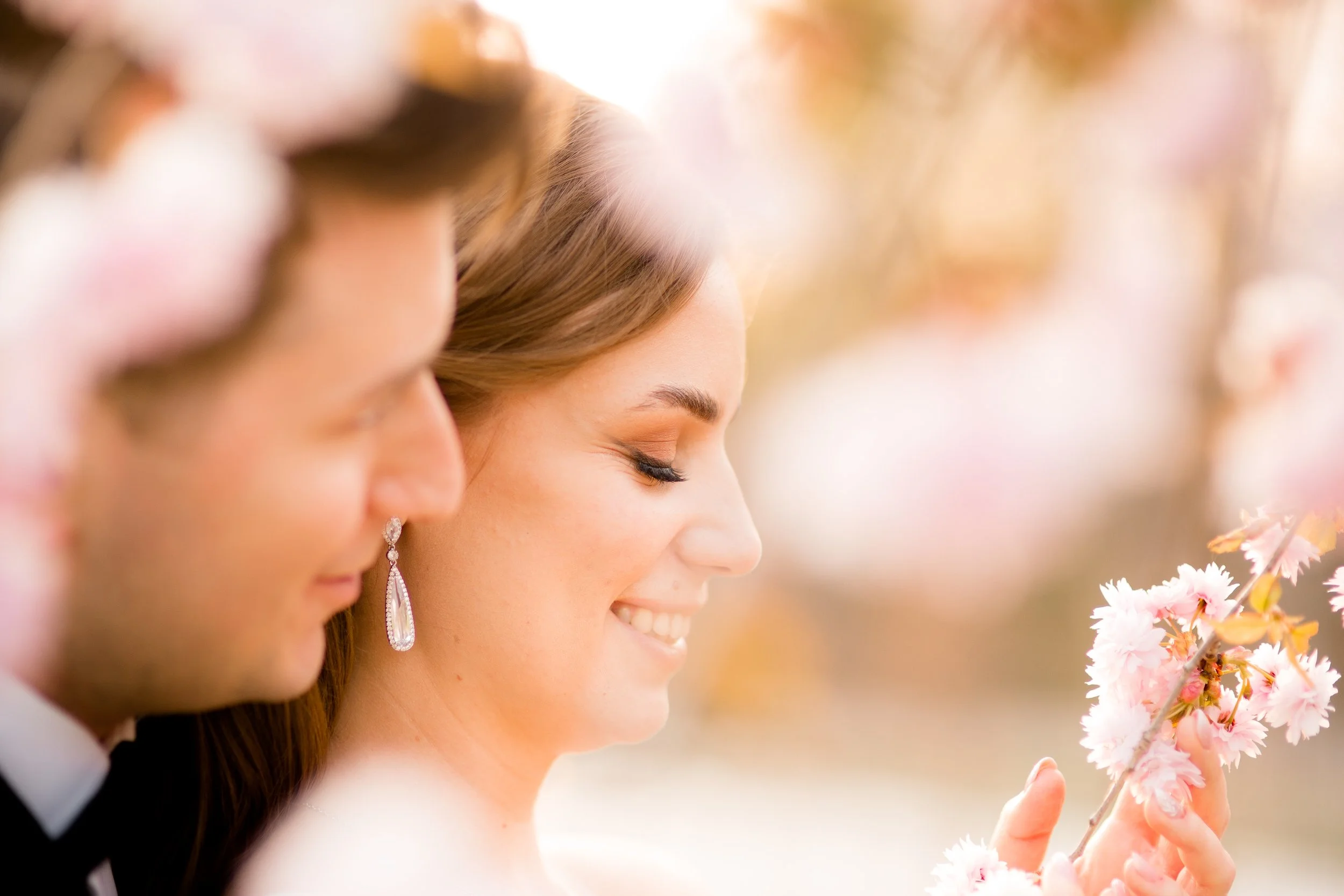 A smiling woman with closed eyes holding pink cherry blossom flowers, standing next to a man in formal attire outdoors during golden hour.