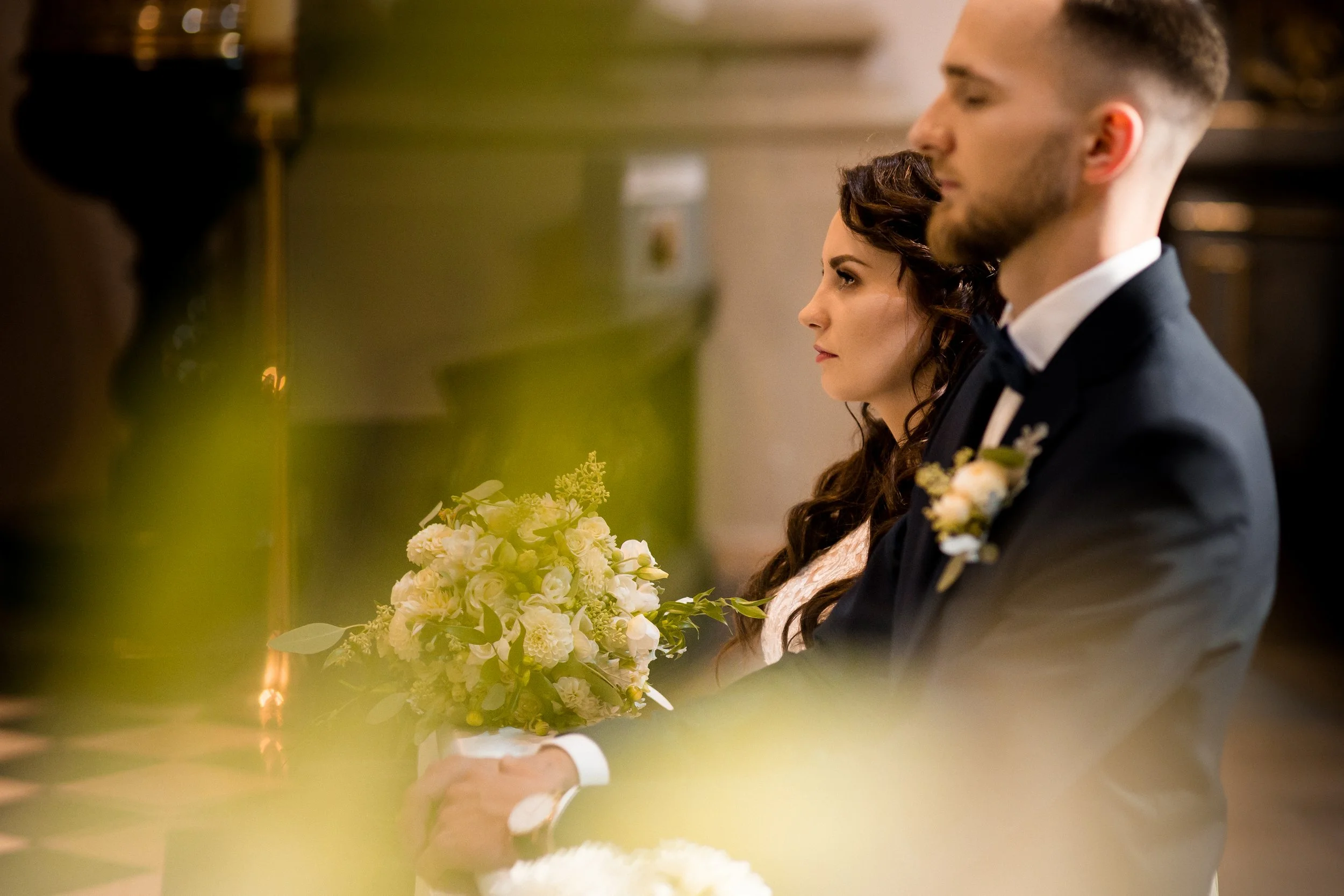 A bride and groom sitting side by side during their wedding ceremony, both with eyes closed and holding hands, with a bouquet of flowers in the bride's lap, inside a church or similar venue.