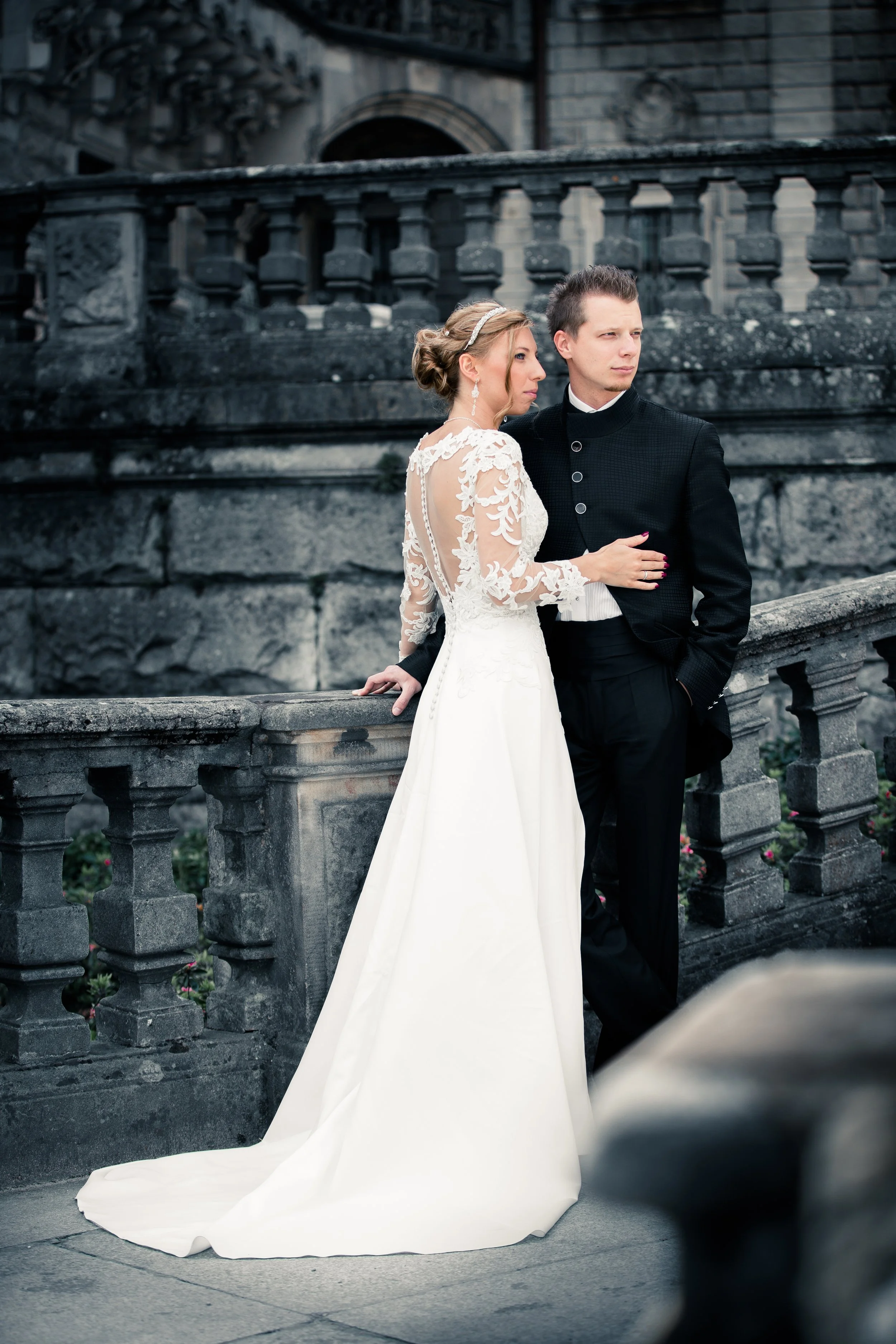 A bride and groom standing close together on a stone bridge, with the bride in a white wedding dress with lace sleeves and the groom in a dark suit, in front of an old stone building.