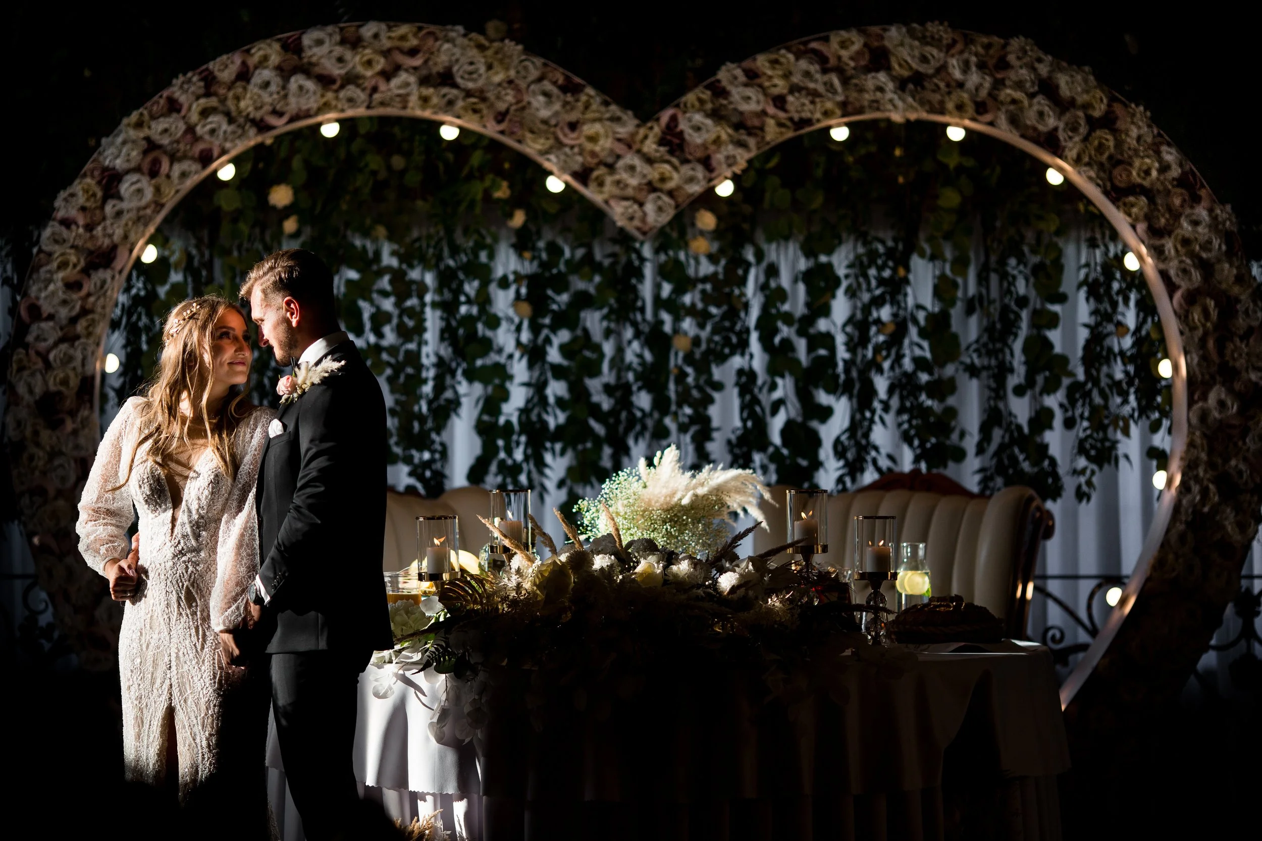 Bride and groom standing close together at their wedding reception, with floral heart-shaped arch decorated with lights in the background and a beautifully arranged table with flowers and candles in front.