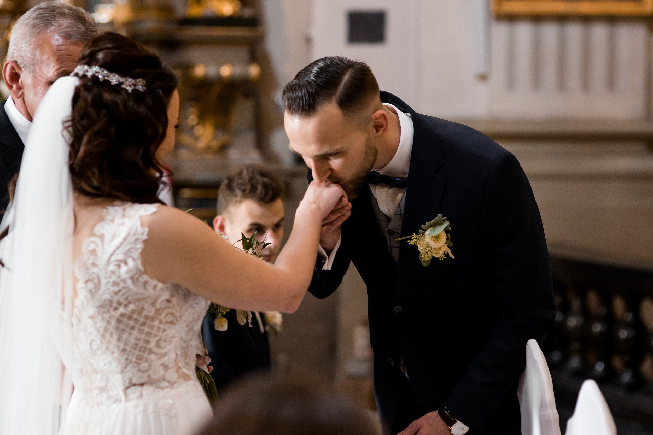 A groom kisses a bride's hand during a wedding ceremony, with a young boy and family members present, in an elegant indoor setting.