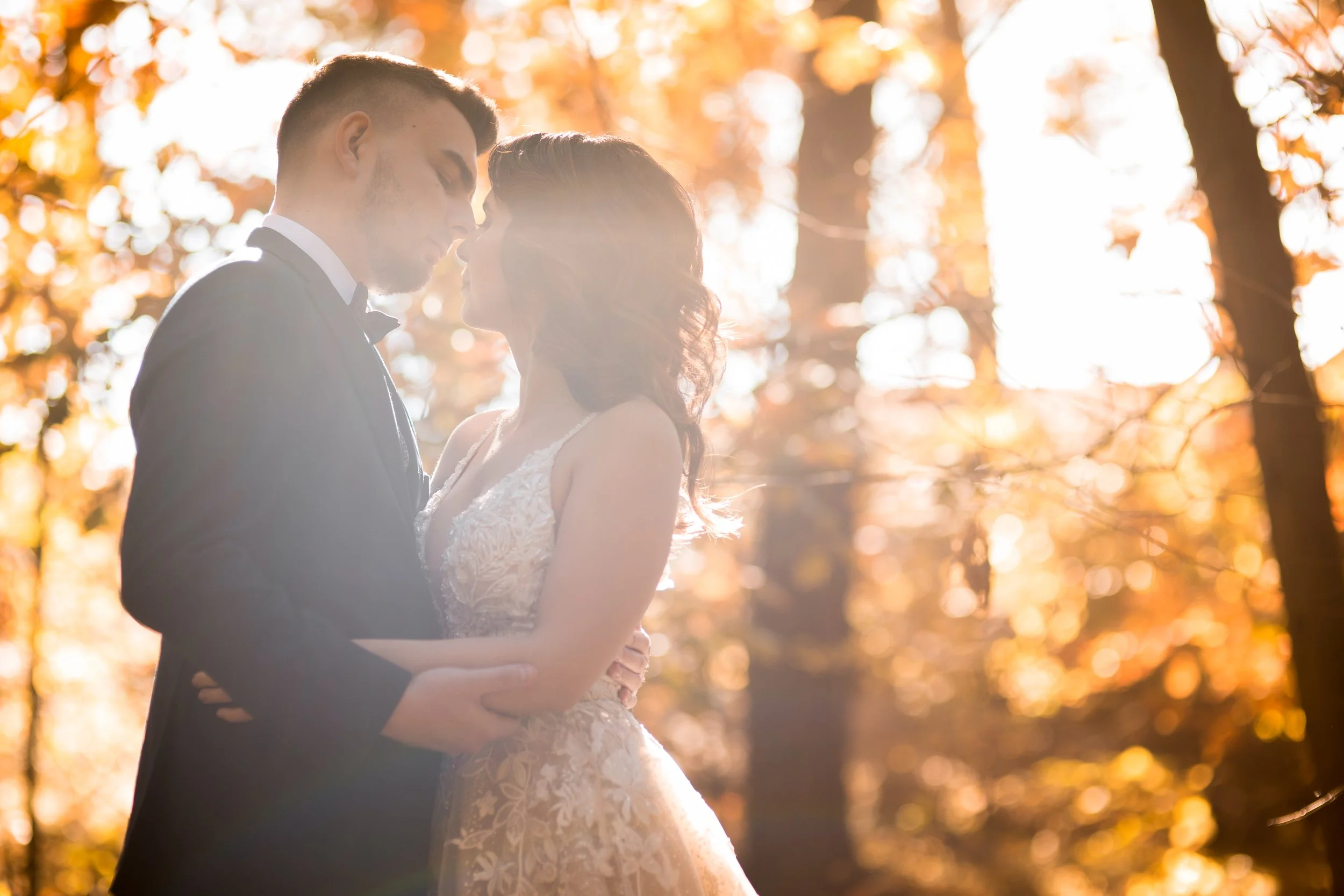 A bride and groom standing close together in a forest during autumn, with the sun shining through orange leaves, sharing an intimate moment.