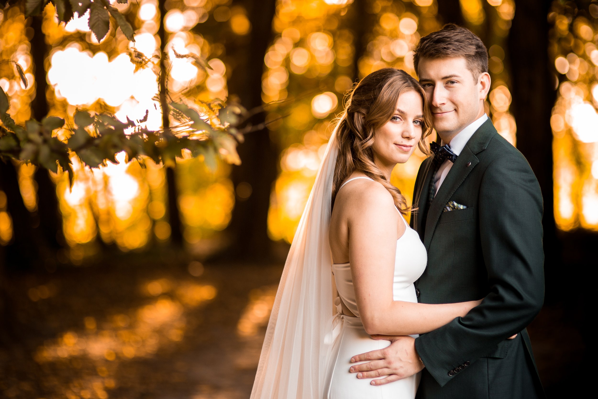 A bride and groom standing close together outdoors during sunset, surrounded by trees with golden leaves in the background.