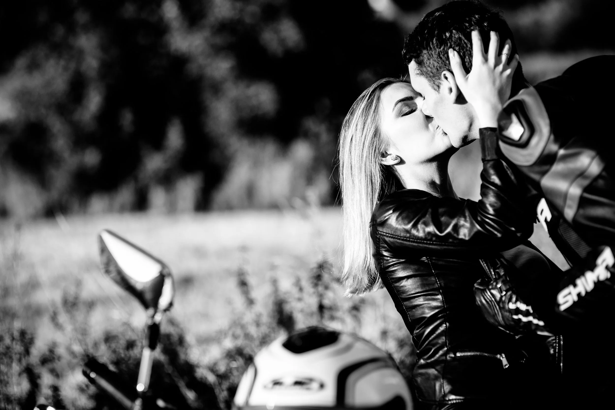 A black and white photo of a couple sharing a kiss outdoors next to a motorcycle, with a scenic, blurred background.