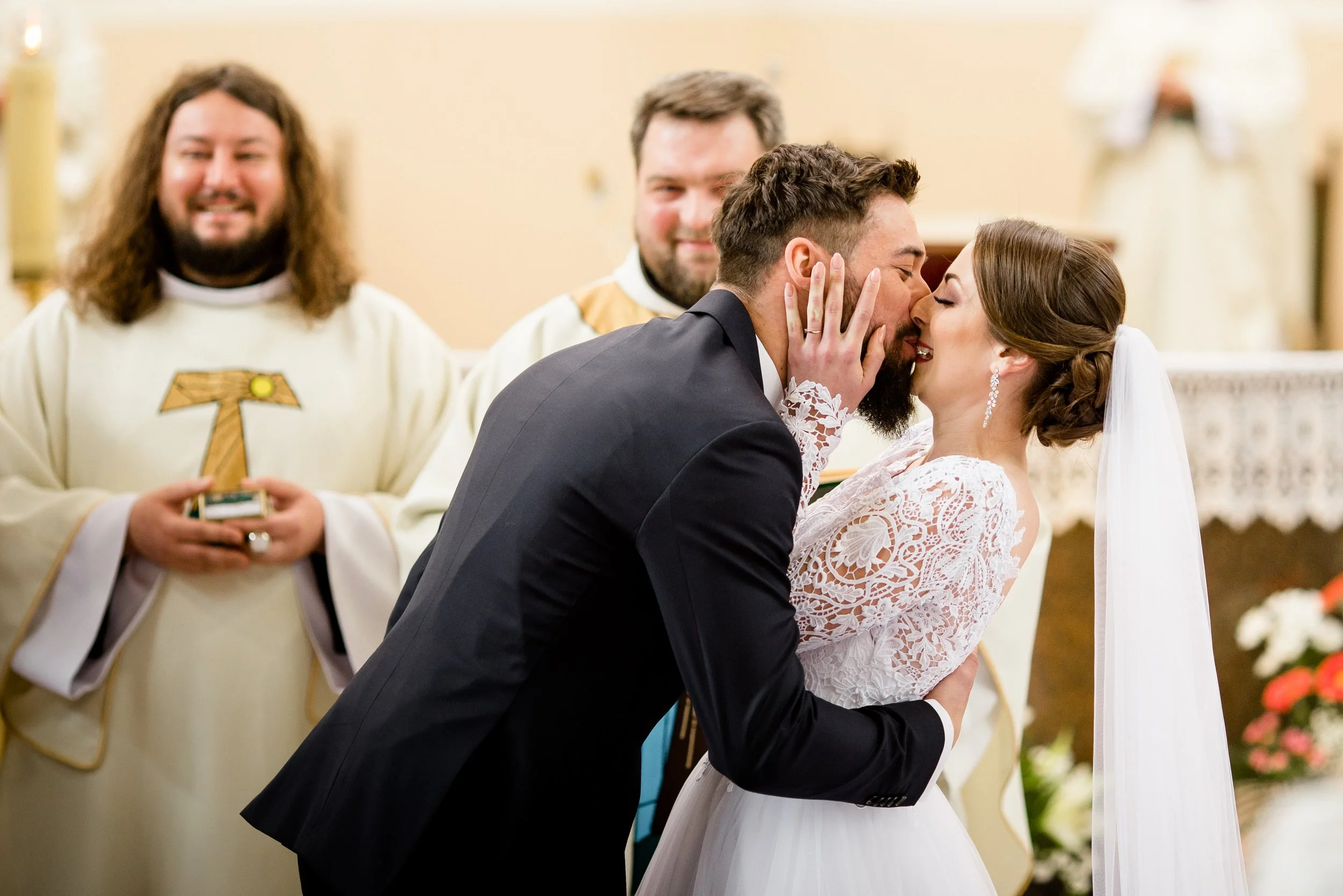 A bride and groom sharing a kiss during their wedding ceremony, with two priests in the background.