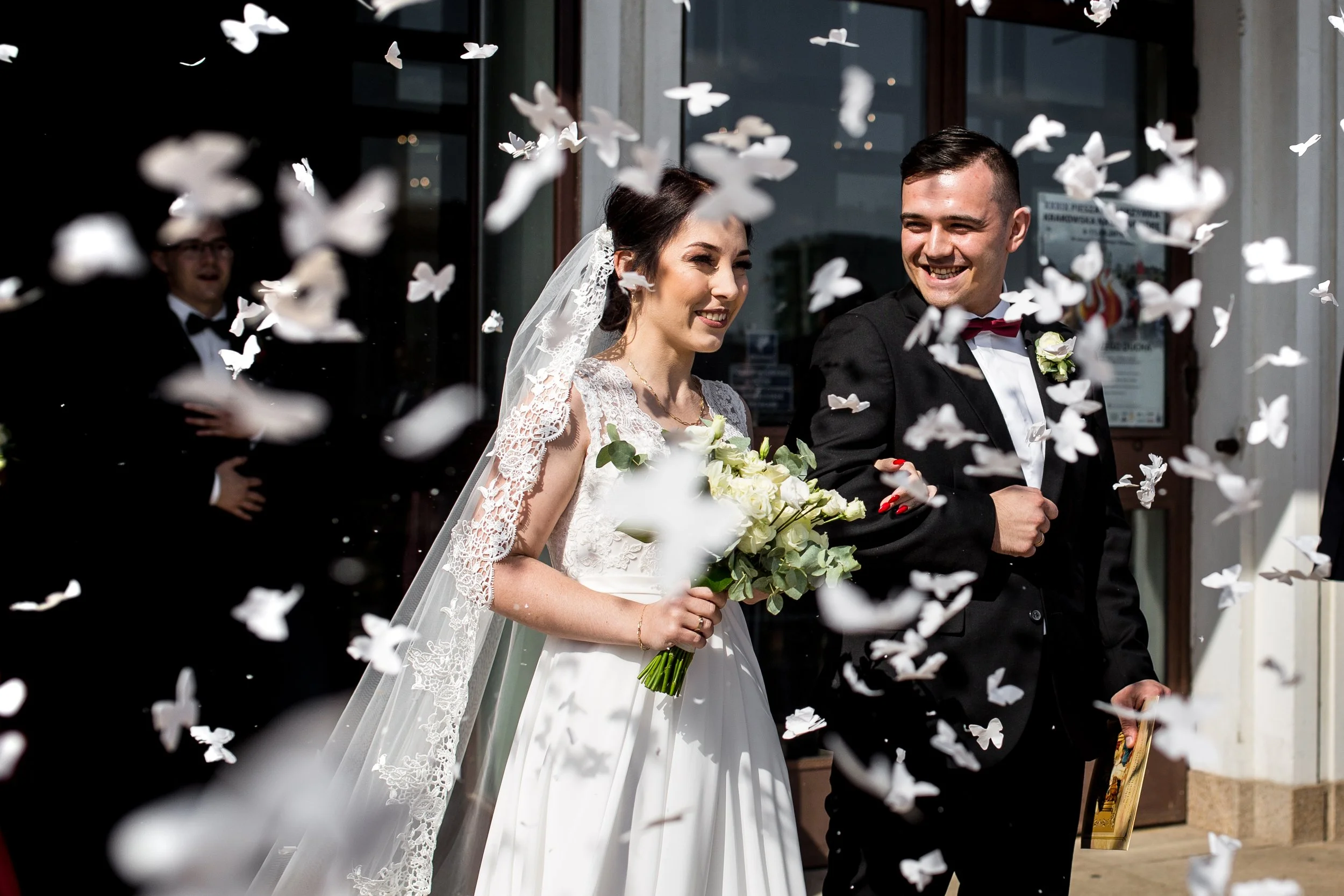 A bride and groom smiling outside a building, surrounded by falling white paper butterflies, celebrating their wedding.