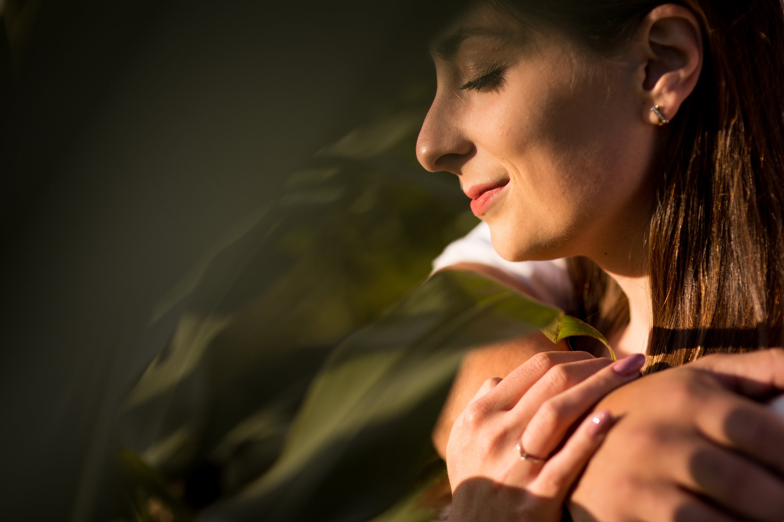 A woman with closed eyes and a gentle smile holds a large green leaf close to her face, in warm, natural lighting.