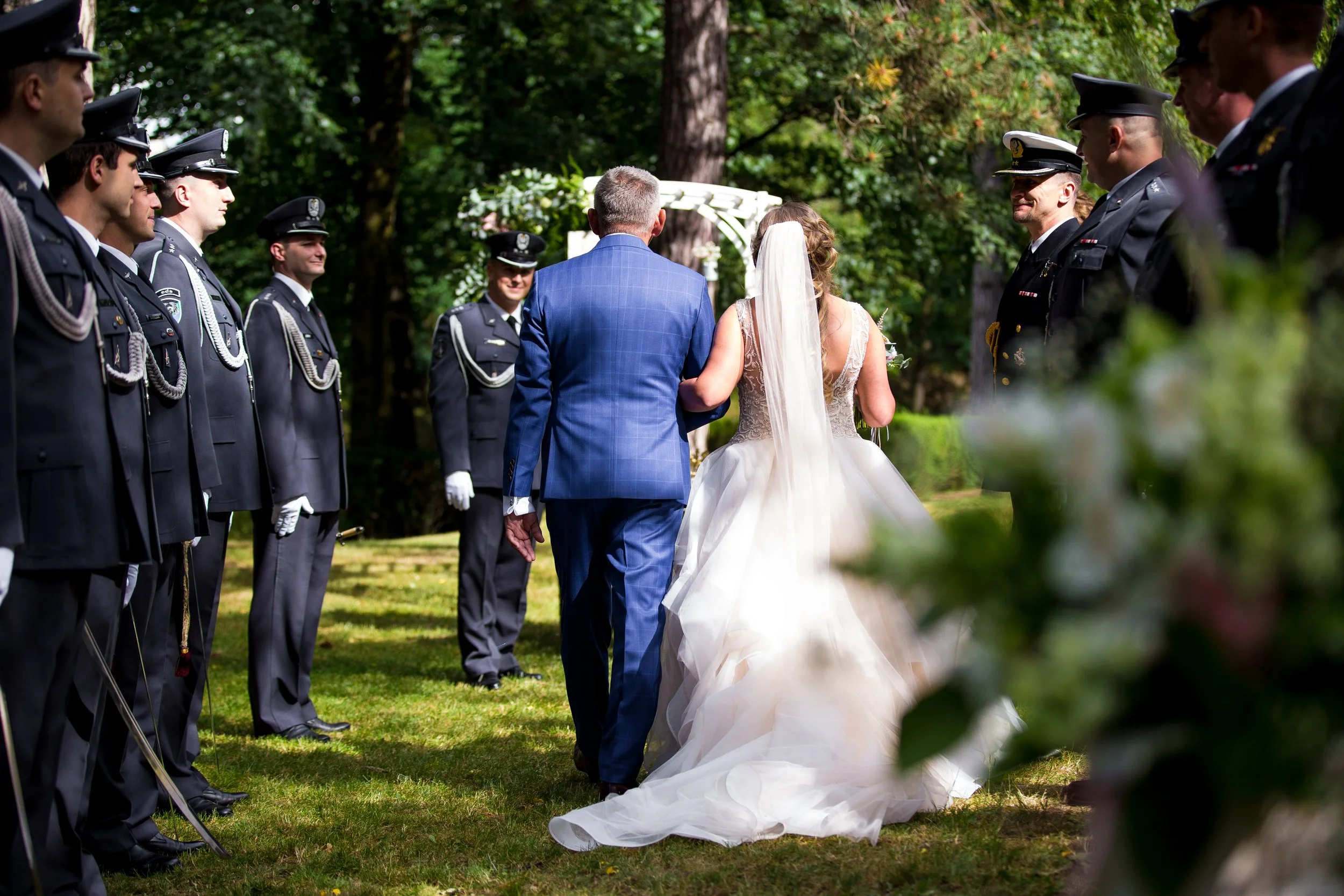 A bride walking with her father towards an outdoor wedding ceremony, with uniformed officers standing on both sides under a canopy in a wooded area.