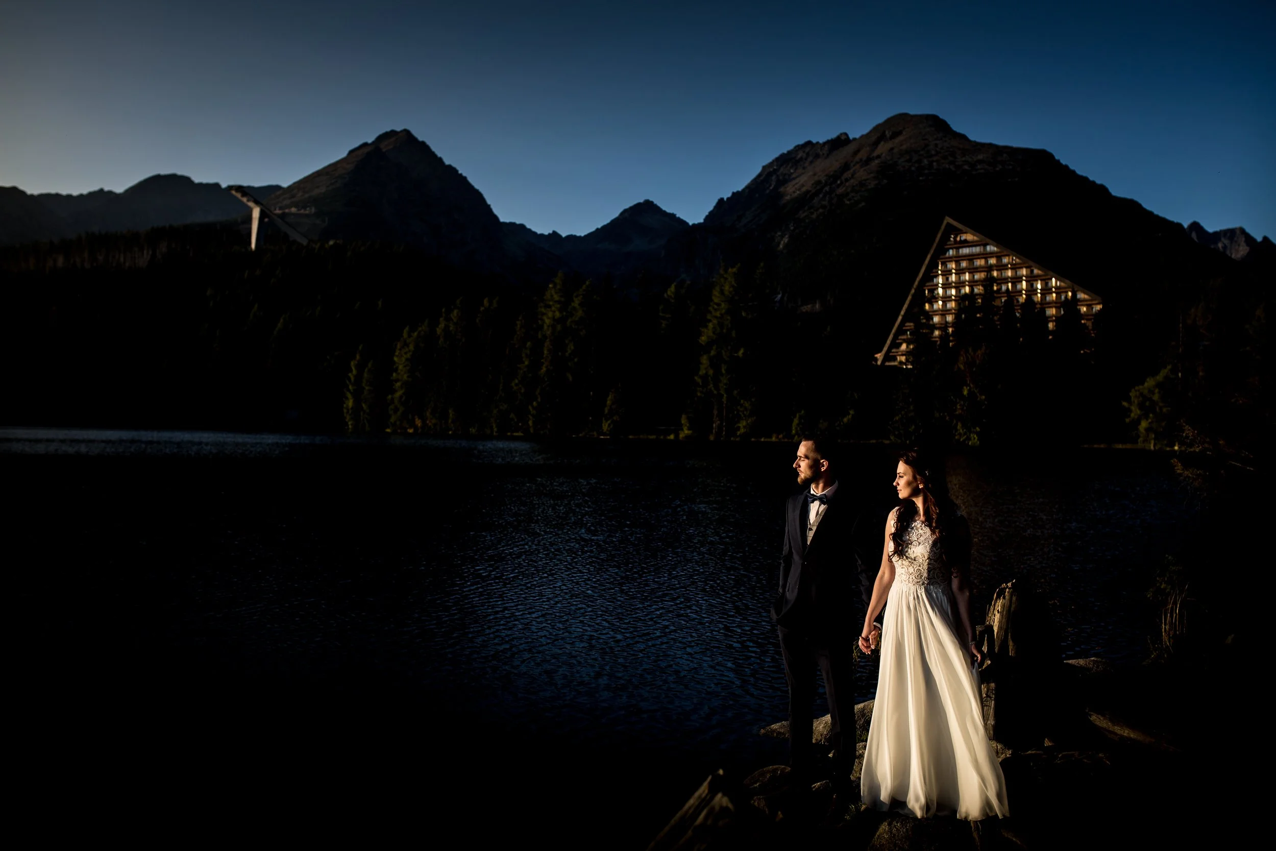 A bride and groom holding hands by a lake at sunset, with mountains and a modern triangular building in the background.