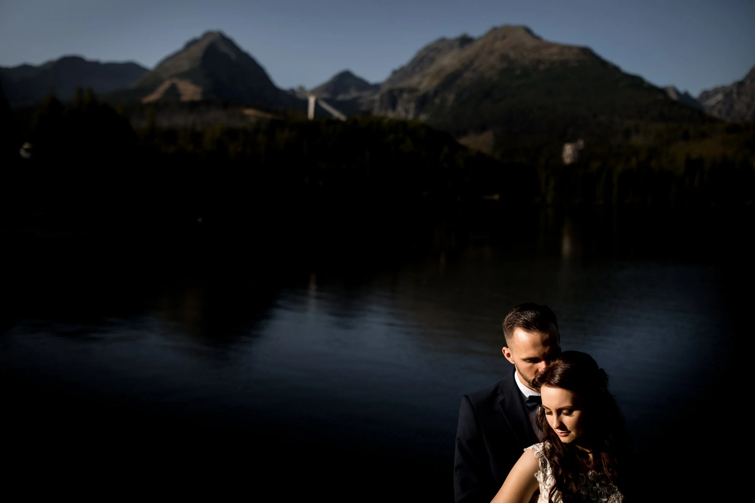 A couple dressed in formal attire, embracing by a darkened body of water with mountains in the background.