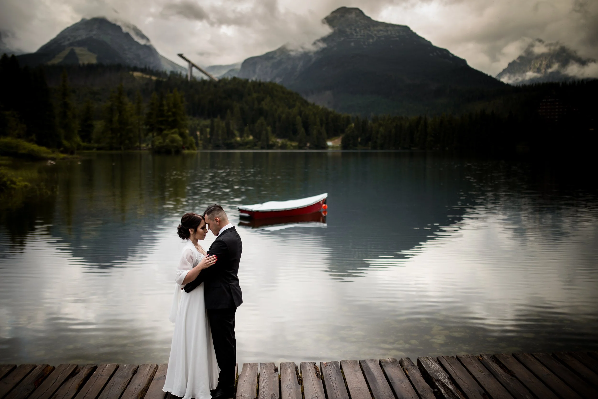 A wedding couple is embracing on a wooden dock by a lake with mountains and dense forest in the background. The woman is wearing a white dress and the man is in a tuxedo. A small red and white boat floats on the water behind them.