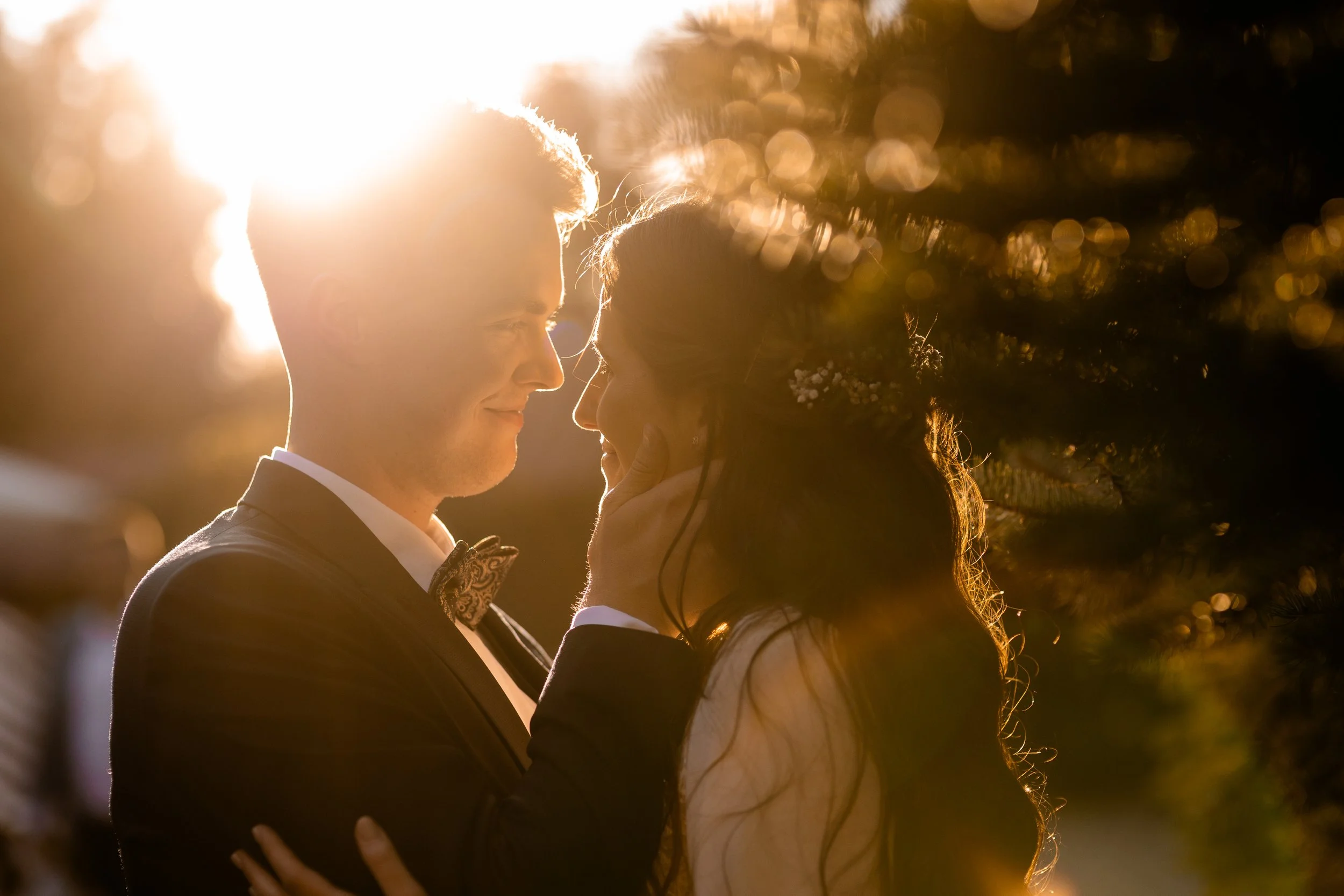 Para w dniu ślubu, stojąca naprzeciwko siebie z uśmiechami na twarzy, na zewnątrz o zachodzie słońca, a promienie słoneczne tworzą wokół nich ciepłą poświatę.