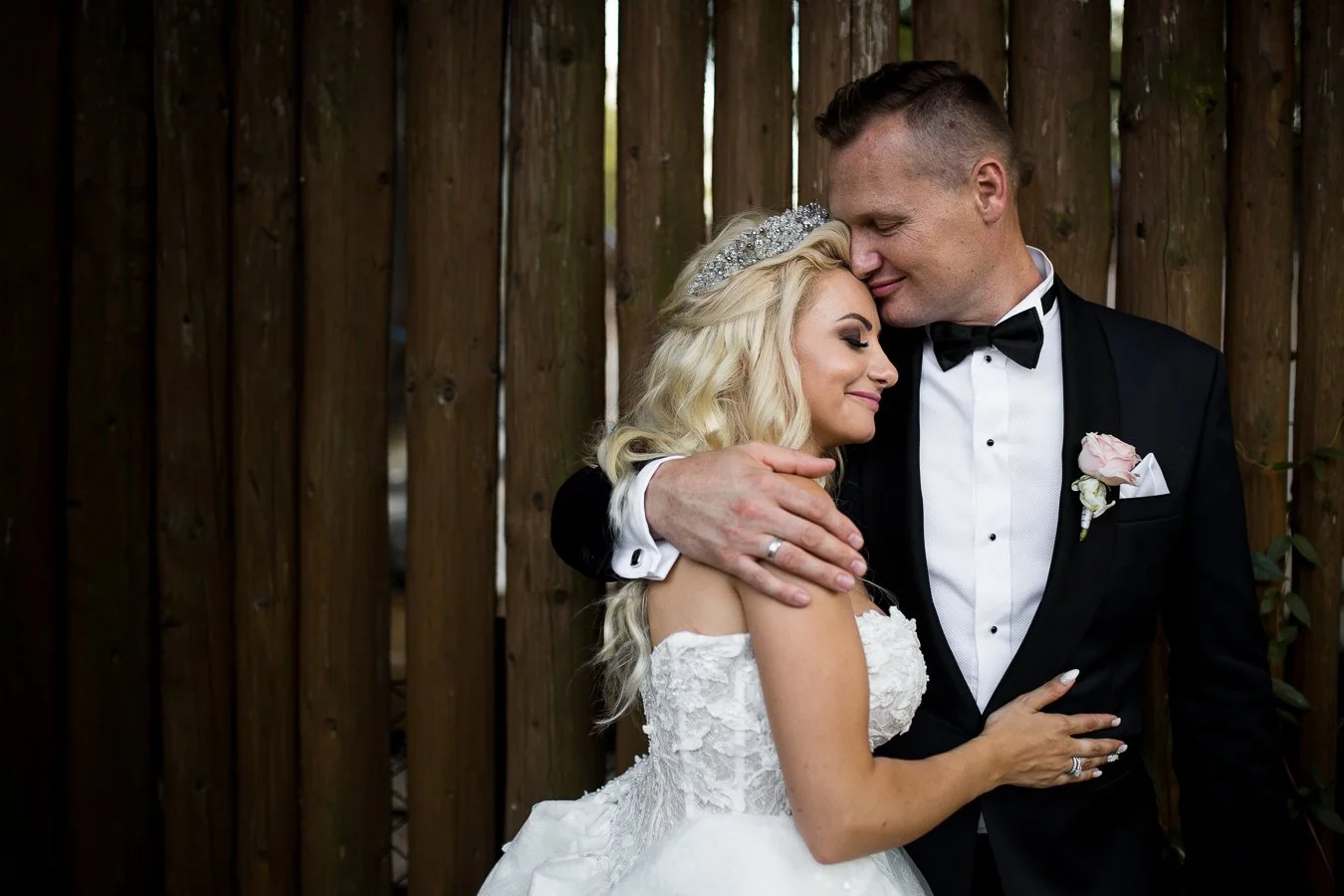 A bride and groom embrace lovingly, smiling with eyes closed, standing in front of a wooden fence during their wedding.