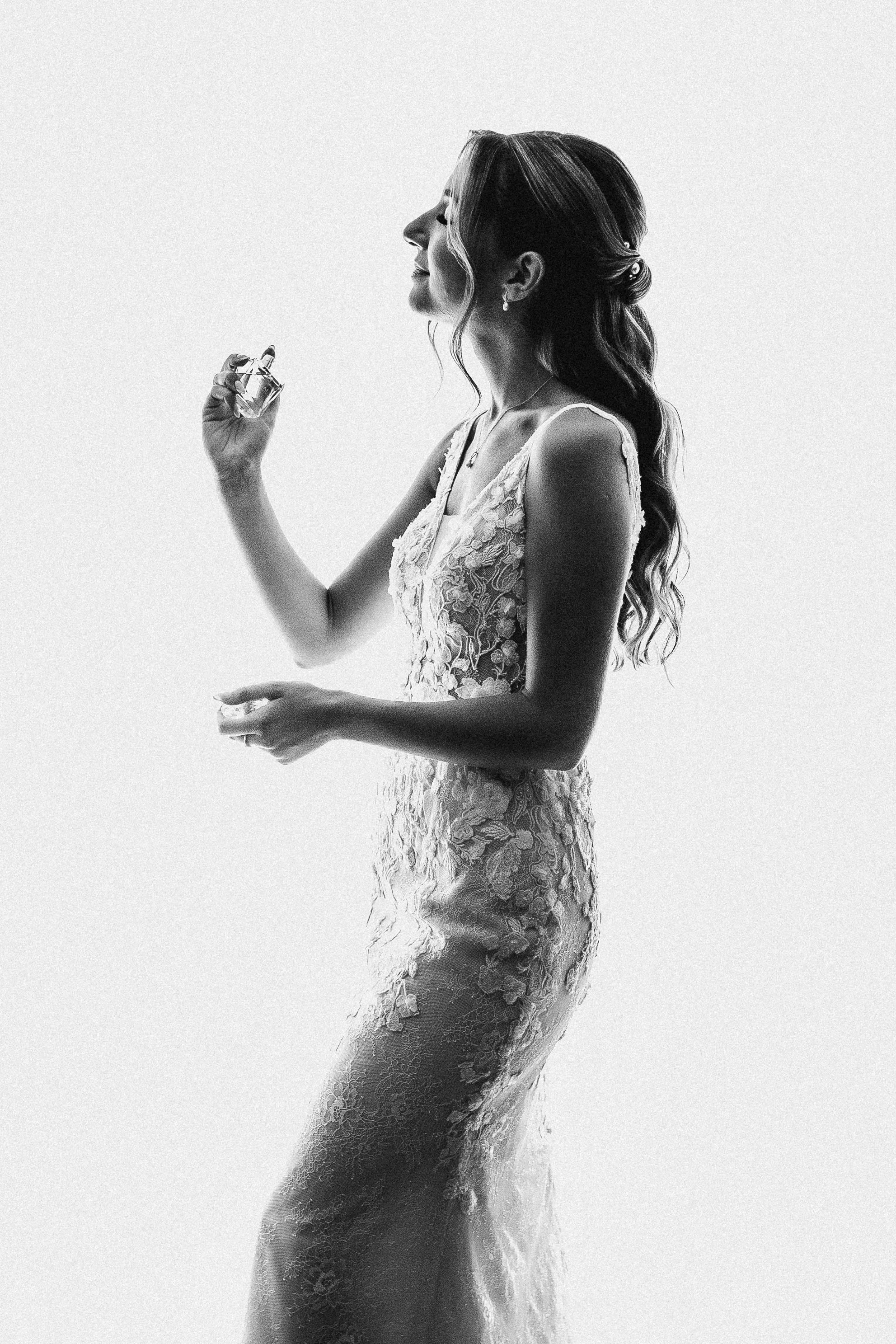 A woman in a wedding dress holding a perfume bottle in her right hand and a small container in her left, smiling with her head tilted back.