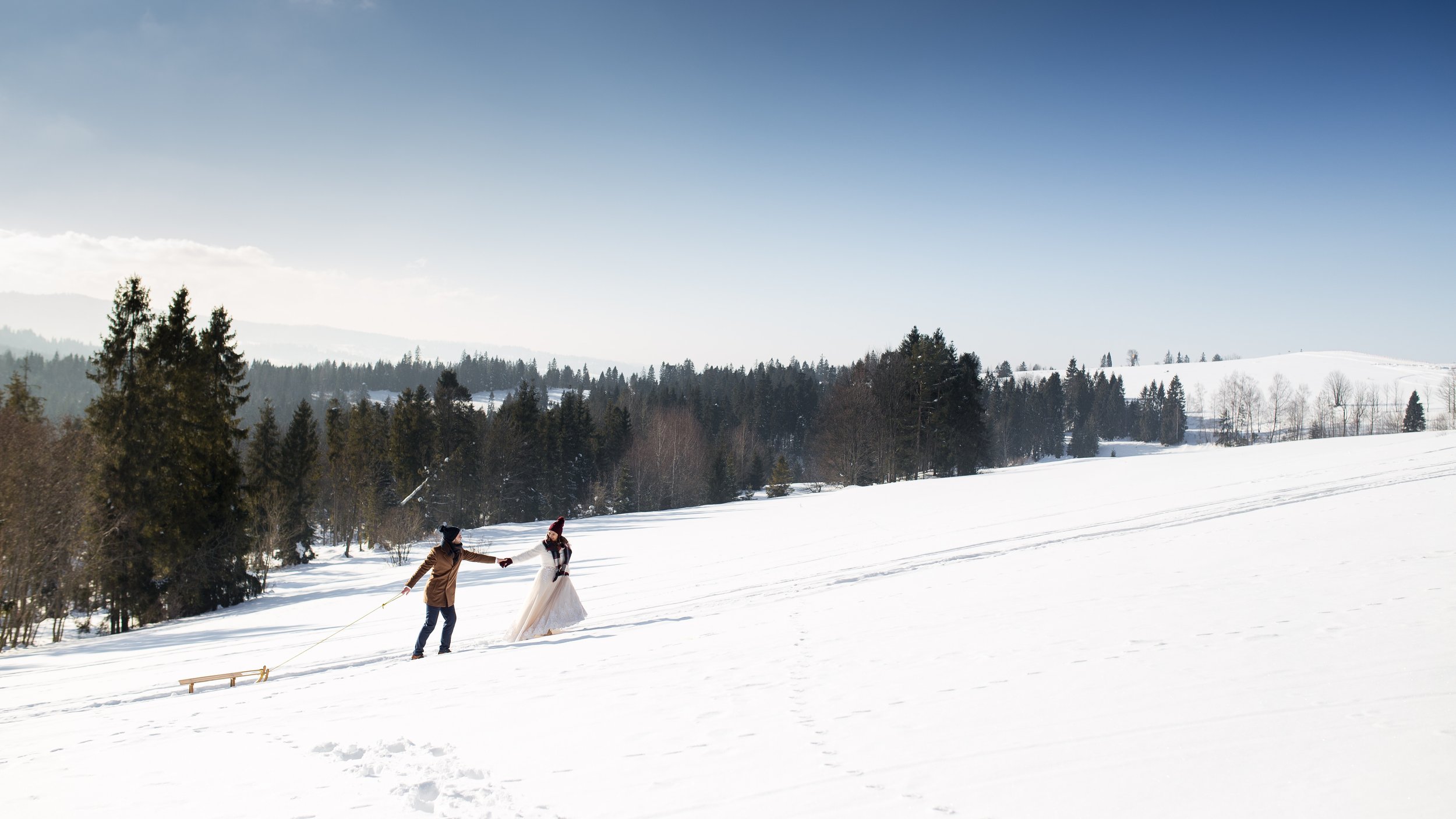 A couple in winter clothing walking through a snowy landscape, with one person pulling a sled, surrounded by trees and distant snow-covered hills under a clear sky.