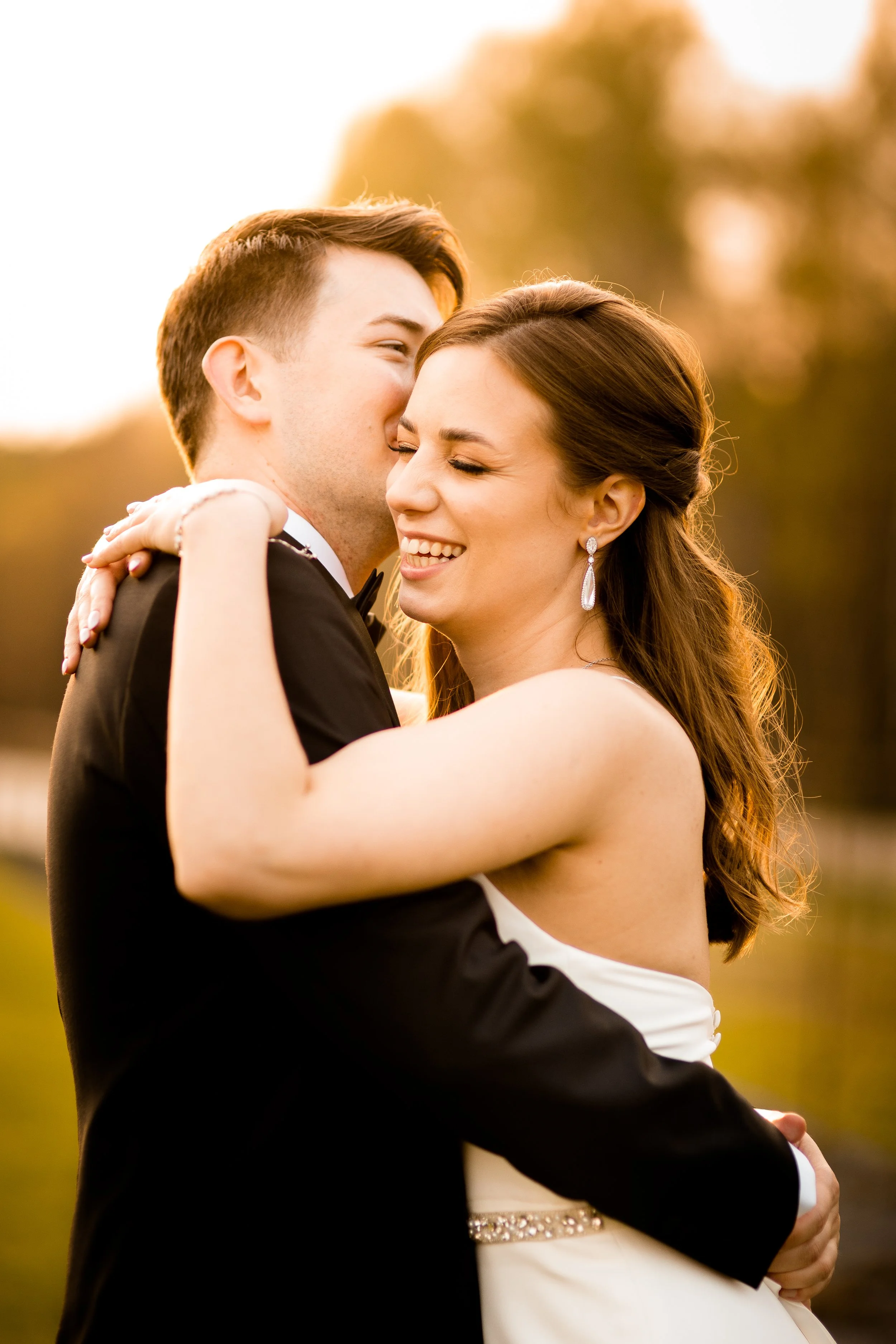 A happy bride and groom sharing a warm embrace outdoors during sunset, both smiling with eyes closed.